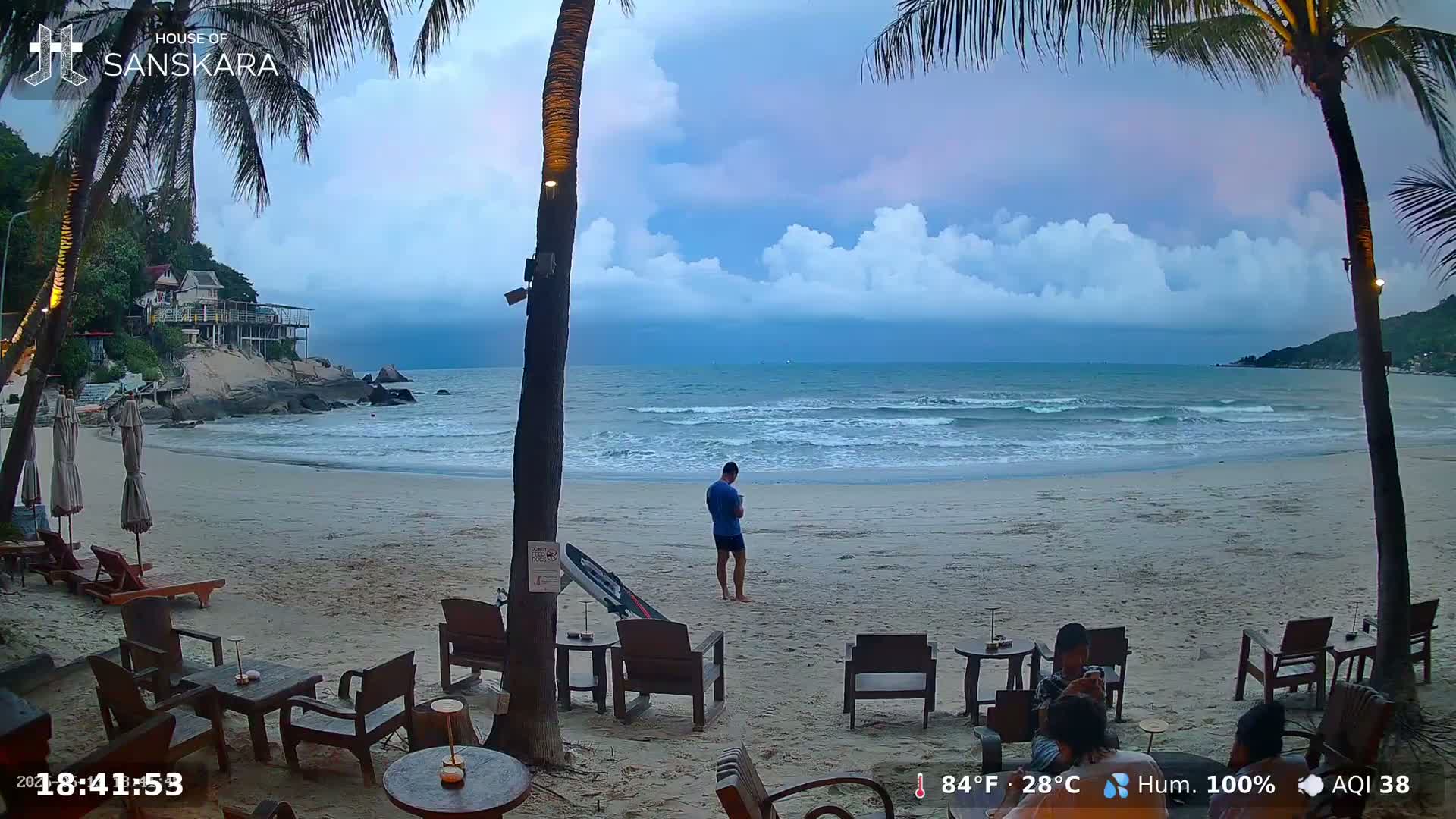 A sandy beach with palm trees, outdoor seating, and a few people, under a partly cloudy sky, with moderate waves on the ocean.