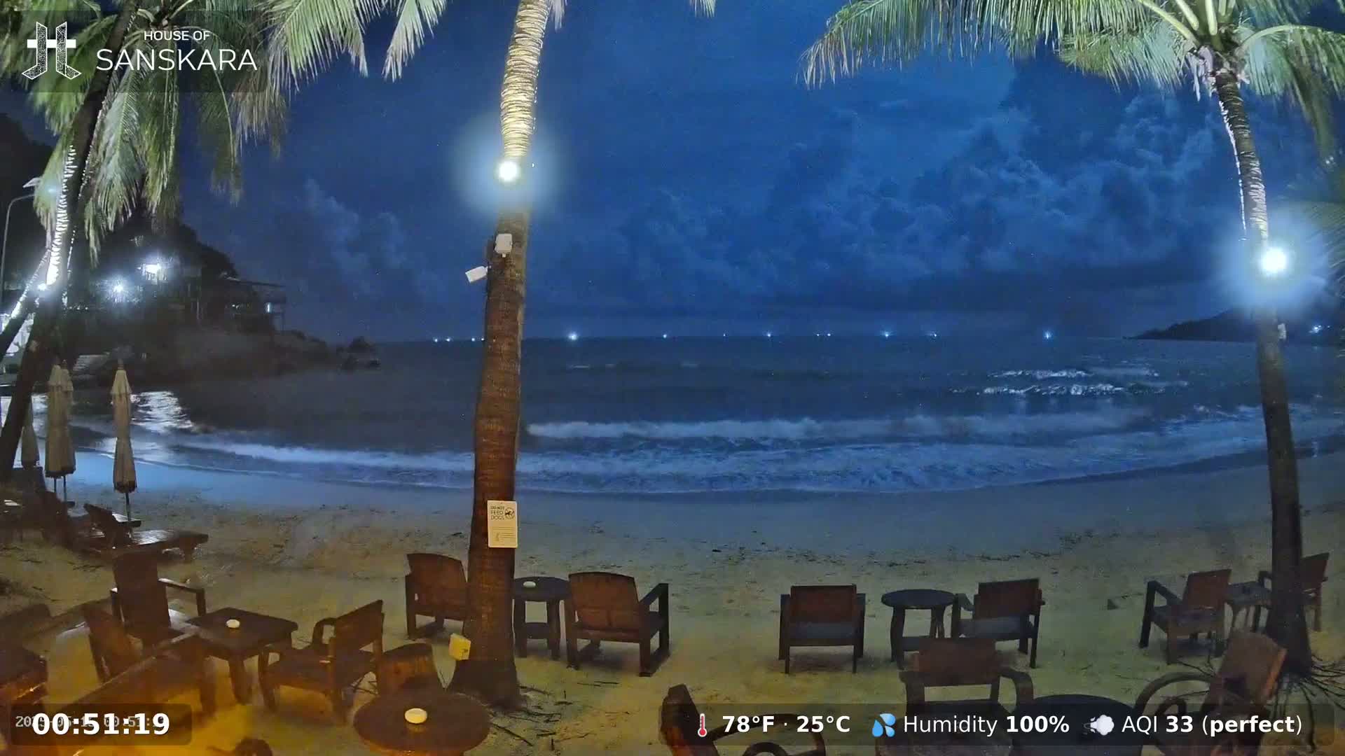 Under a dark, cloudy night sky, a beach with gently lapping waves is shown, featuring empty outdoor seating near palm trees.