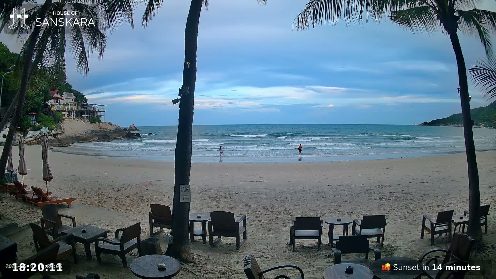 A sandy beach with gentle waves, palm trees, and outdoor seating under a partly cloudy twilight sky.