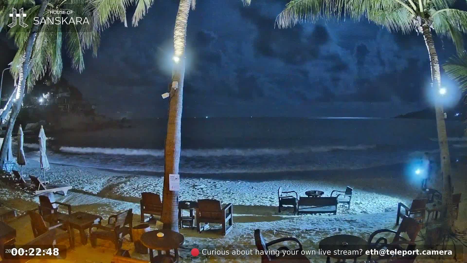 Under a dark, cloudy night sky, gentle waves lap a sandy beach at a beachfront establishment with outdoor seating.