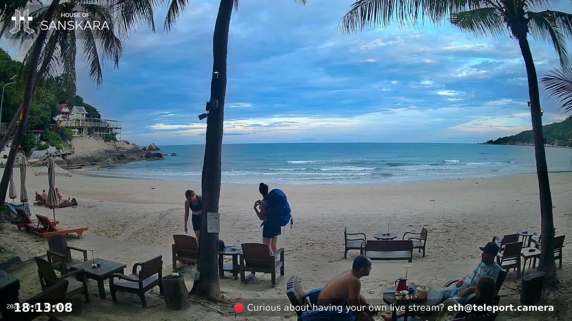 A sandy beach with calm ocean waves under a partly cloudy twilight sky, palm trees, outdoor furniture, and several people are present.