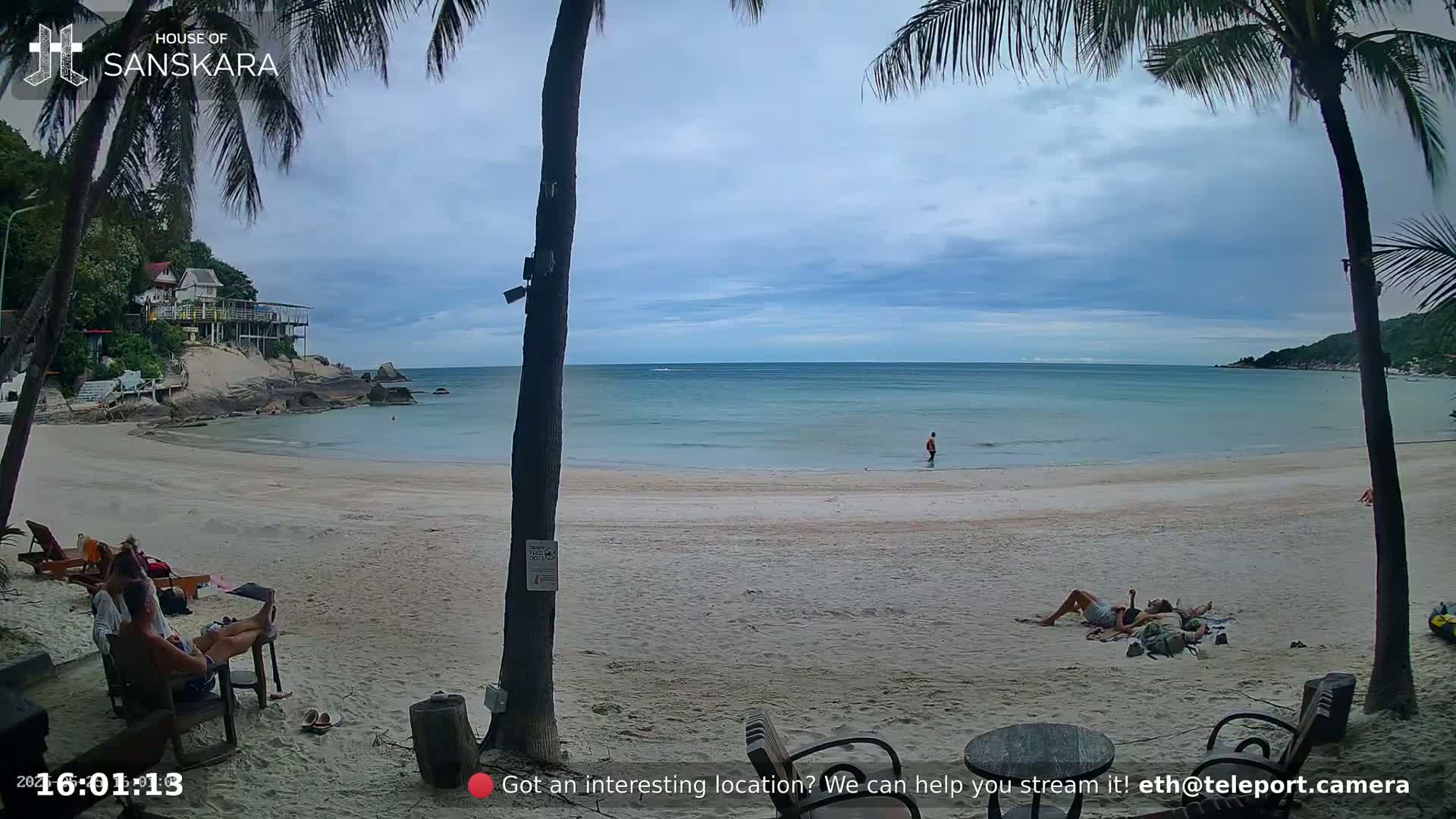 A sandy beach with calm, light blue water, palm trees, and a few people relaxing on the sand under a partly cloudy sky.