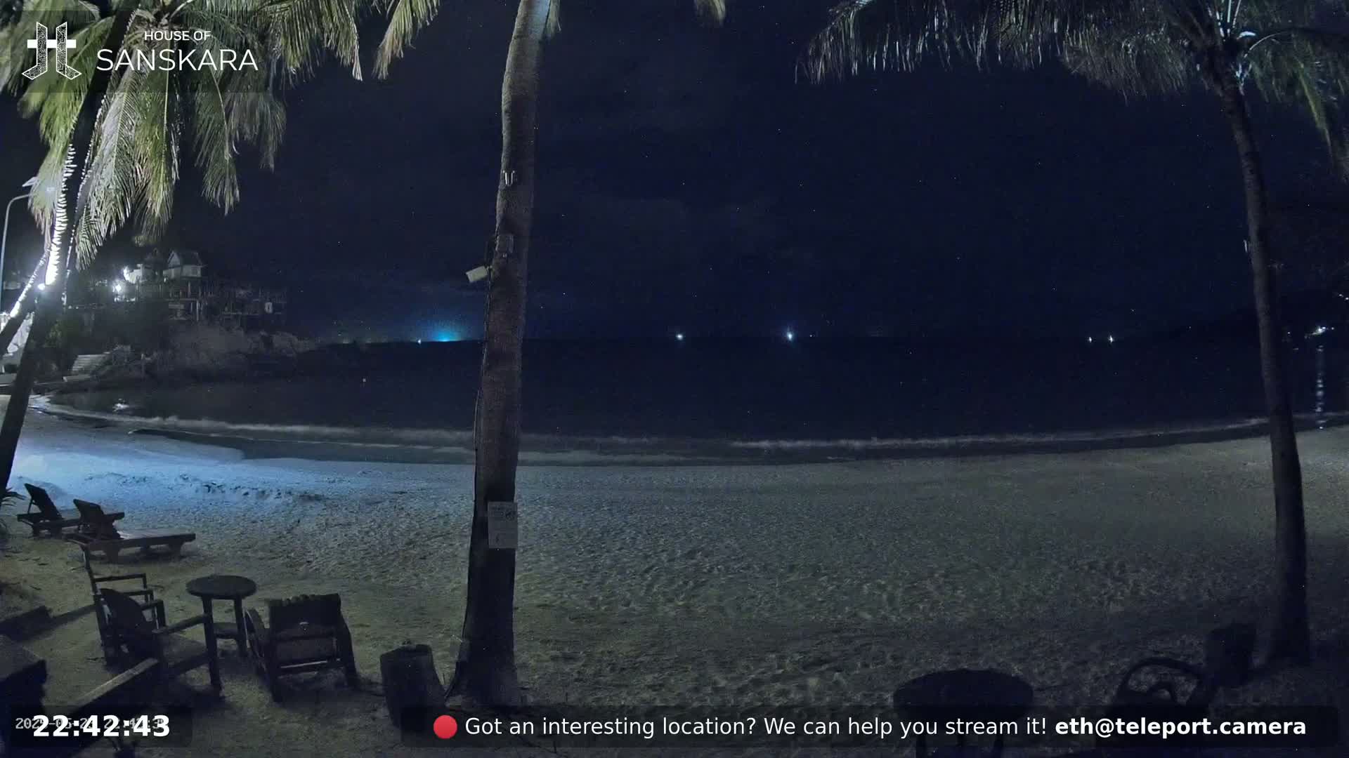 A nighttime beach scene shows a sandy shore with some patio furniture under palm trees, a dark ocean with faint lights in the distance, and a starry sky.
