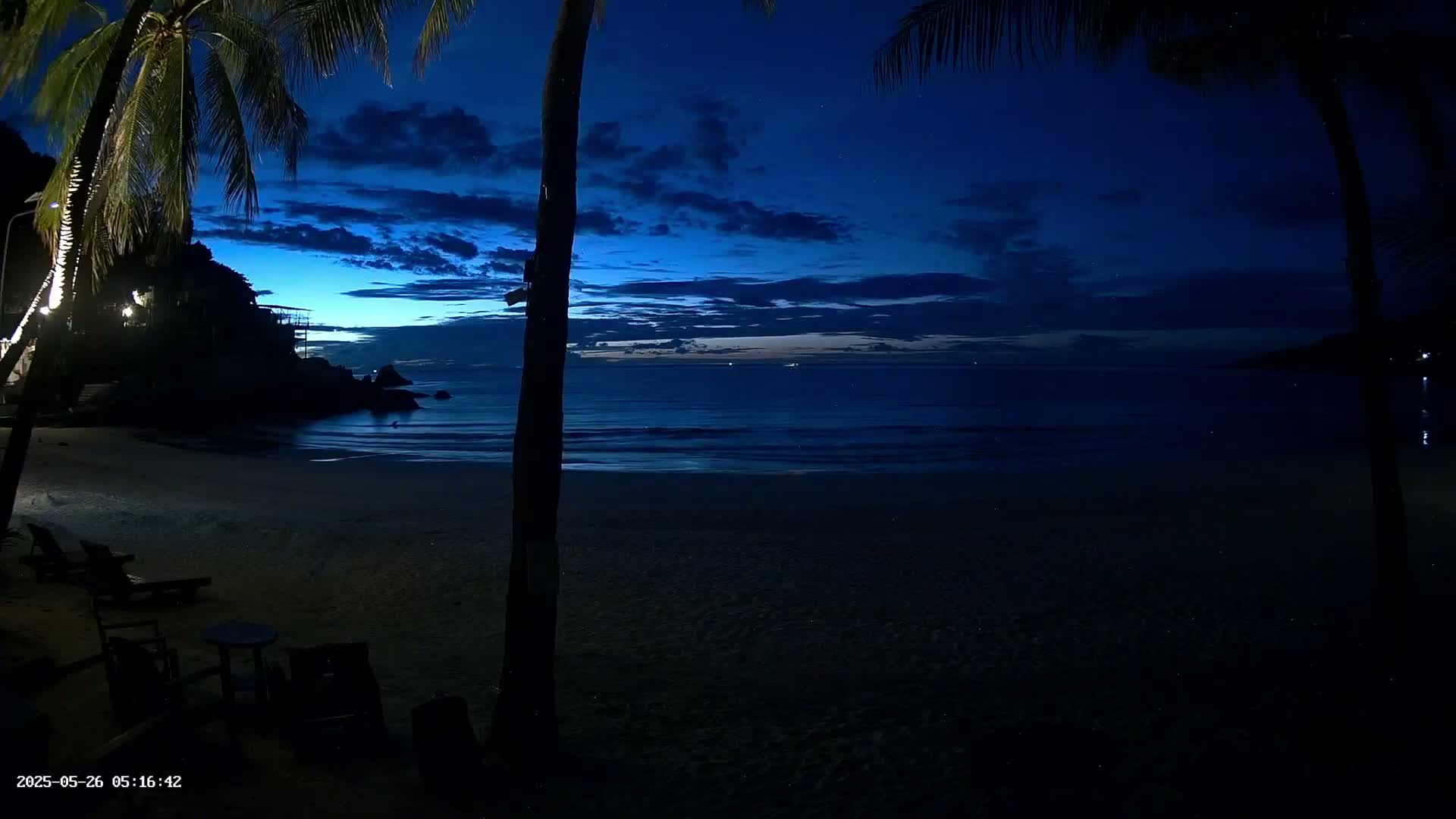 A dark blue twilight sky over a mostly calm ocean, a sandy beach with a few lounge chairs, and palm trees silhouetted against the water.