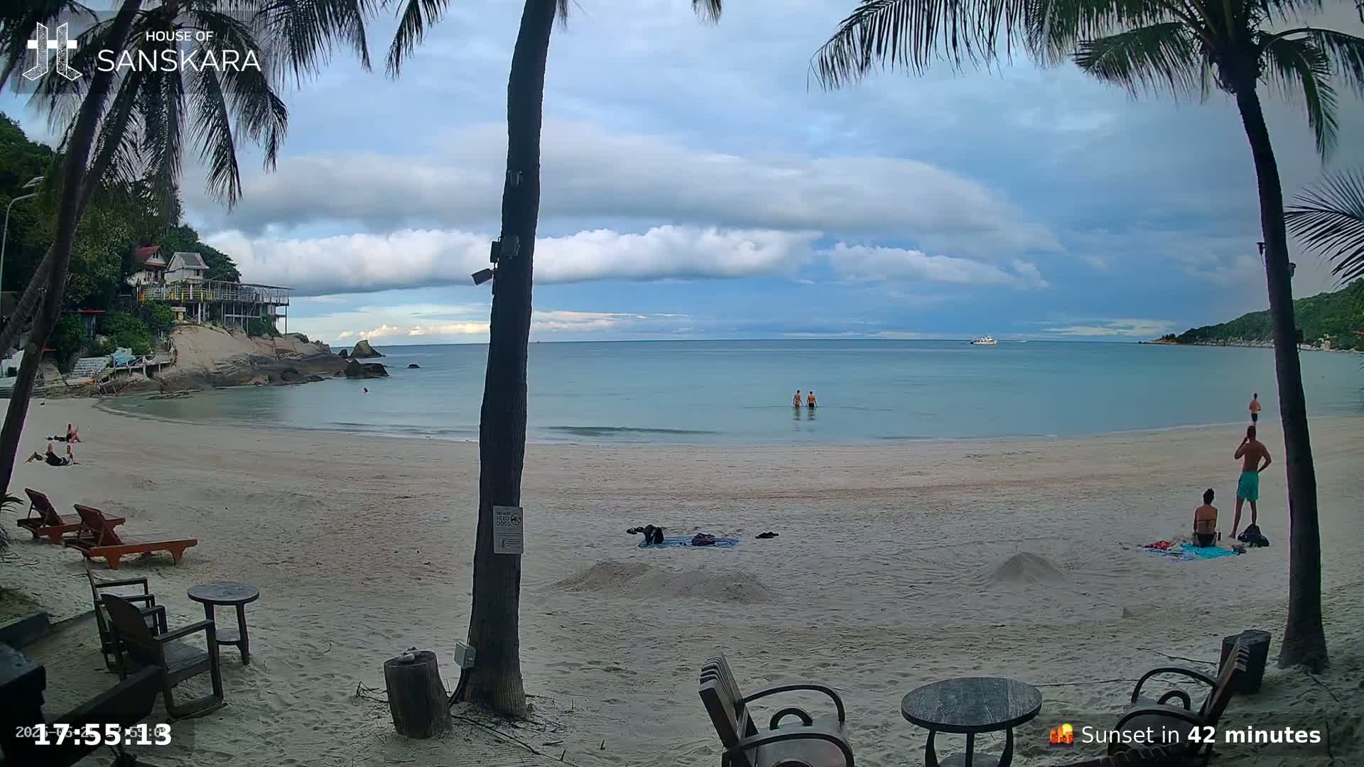 A sandy beach with calm, light-blue water under a partly cloudy sky shows several people relaxing on the sand and in the water, with palm trees and beach furniture in the foreground.