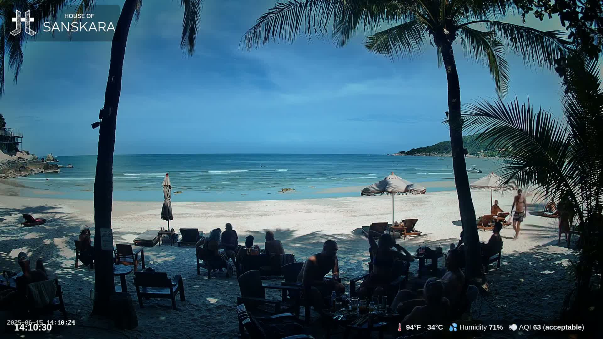 A sunny beach scene shows people relaxing at outdoor tables under palm trees, with calm ocean water in the background.