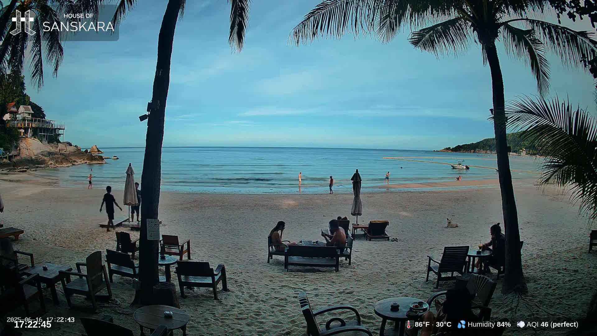 A sandy beach with calm ocean water under a mostly clear sky, palm trees, and several people sitting at outdoor tables.