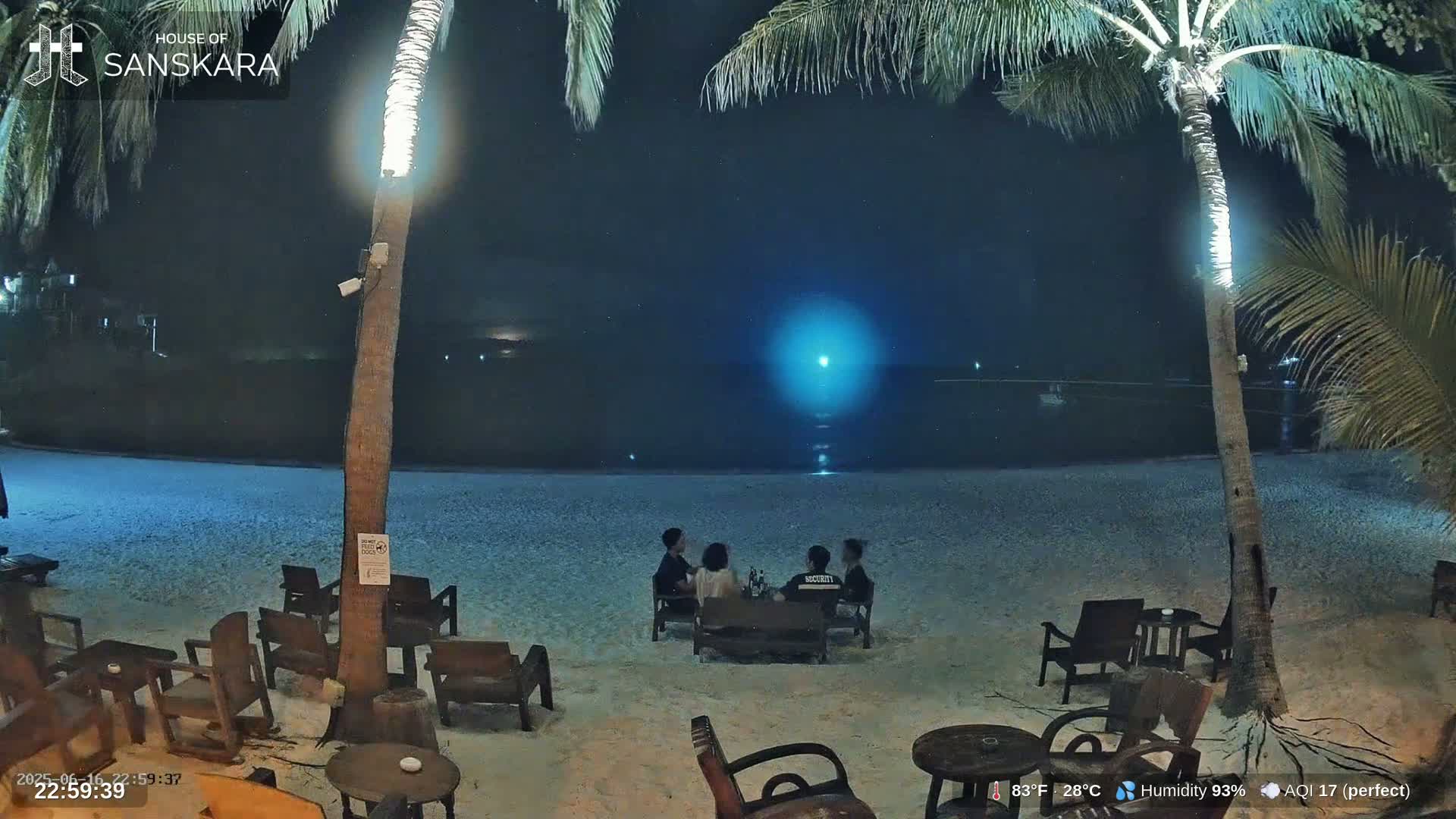 A group of people sits at a table on a sandy beach at night under a partly cloudy sky with a bright light in the distance.