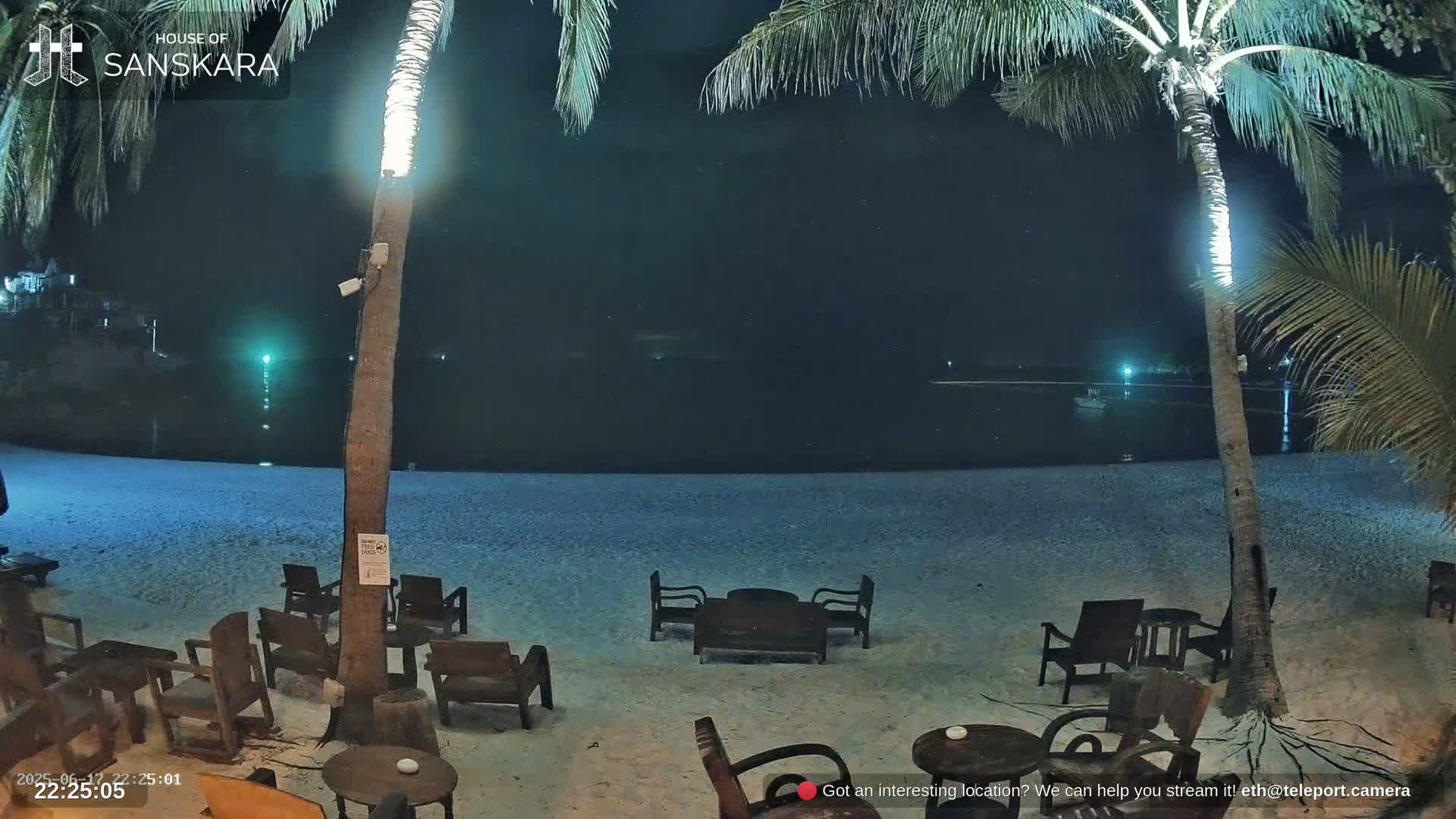 A nighttime beach scene shows several empty wooden chairs and tables on a sandy beach, illuminated by lights on palm trees, with calm dark water in the background.