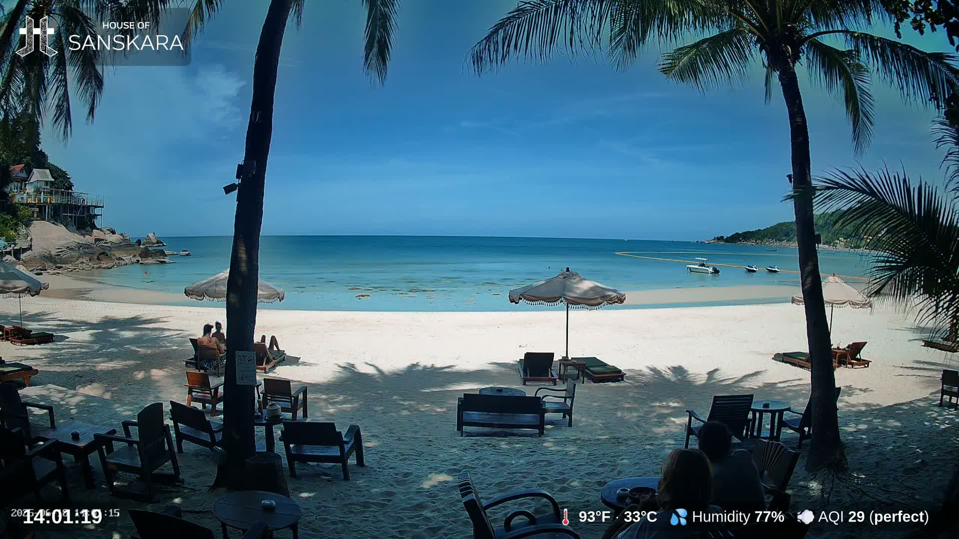 A sunny beach scene with calm, clear turquoise water, palm trees, beach chairs, umbrellas, and a few people relaxing.