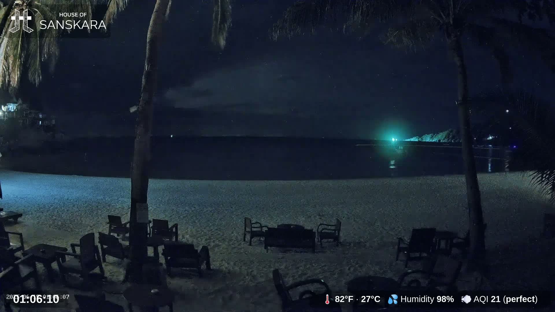 A nighttime beach scene shows empty chairs and tables on a sandy beach, overlooking a dark ocean under a partly cloudy, starry sky.