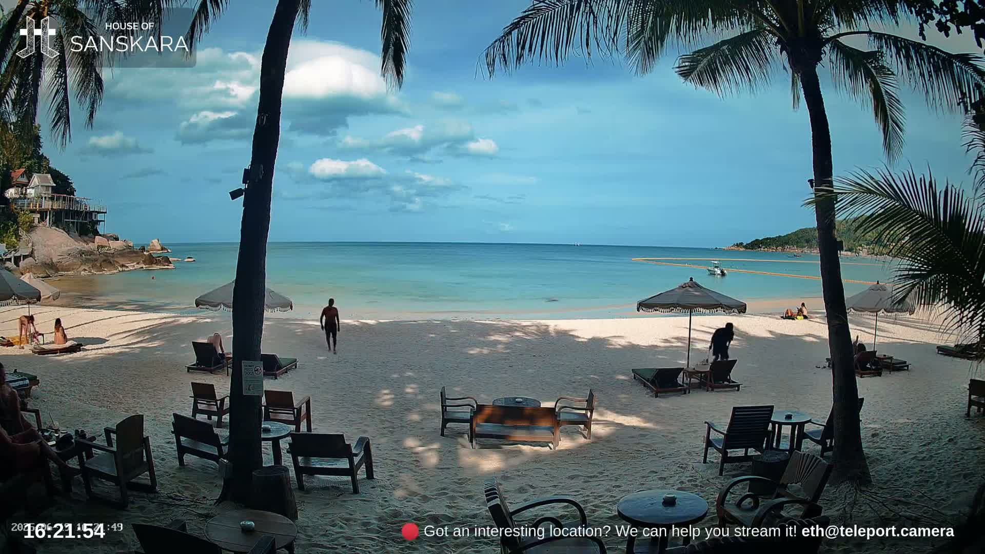 A partly sunny beach scene shows a sandy shore with lounge chairs and umbrellas, palm trees, a calm ocean, and a few people relaxing or walking.