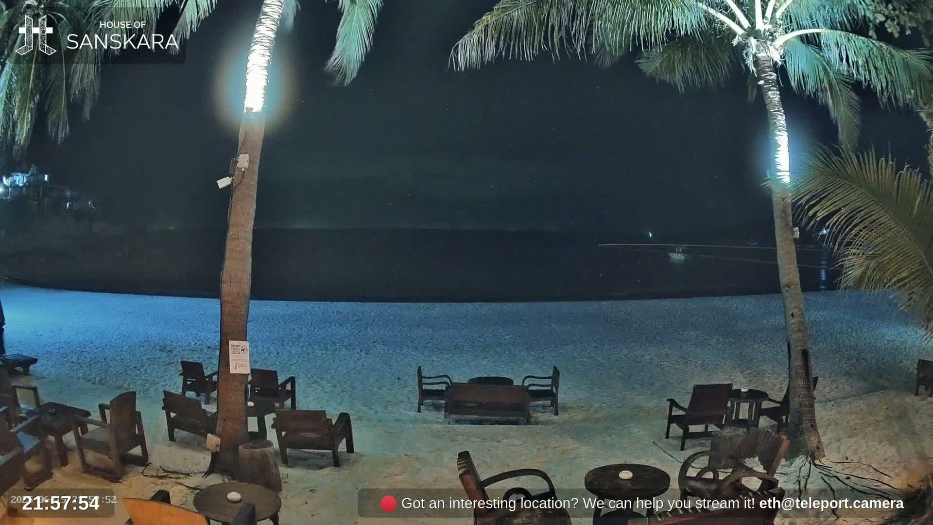 A nighttime beach scene shows empty wooden chairs and tables on a sandy beach under palm trees illuminated by lights, with a dark ocean in the background.