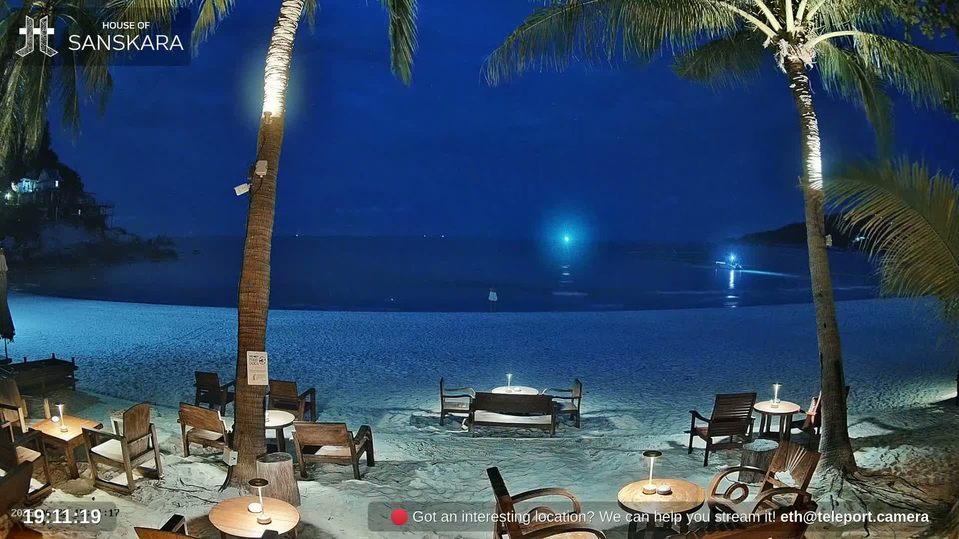 An outdoor nighttime scene shows a beach with palm trees, wooden tables and chairs, and a calm ocean under a dark blue sky.