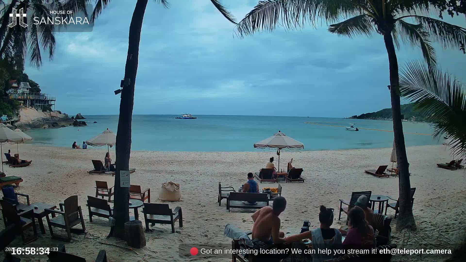A sandy beach with turquoise water, palm trees, beach umbrellas, and several people relaxing under an overcast sky.