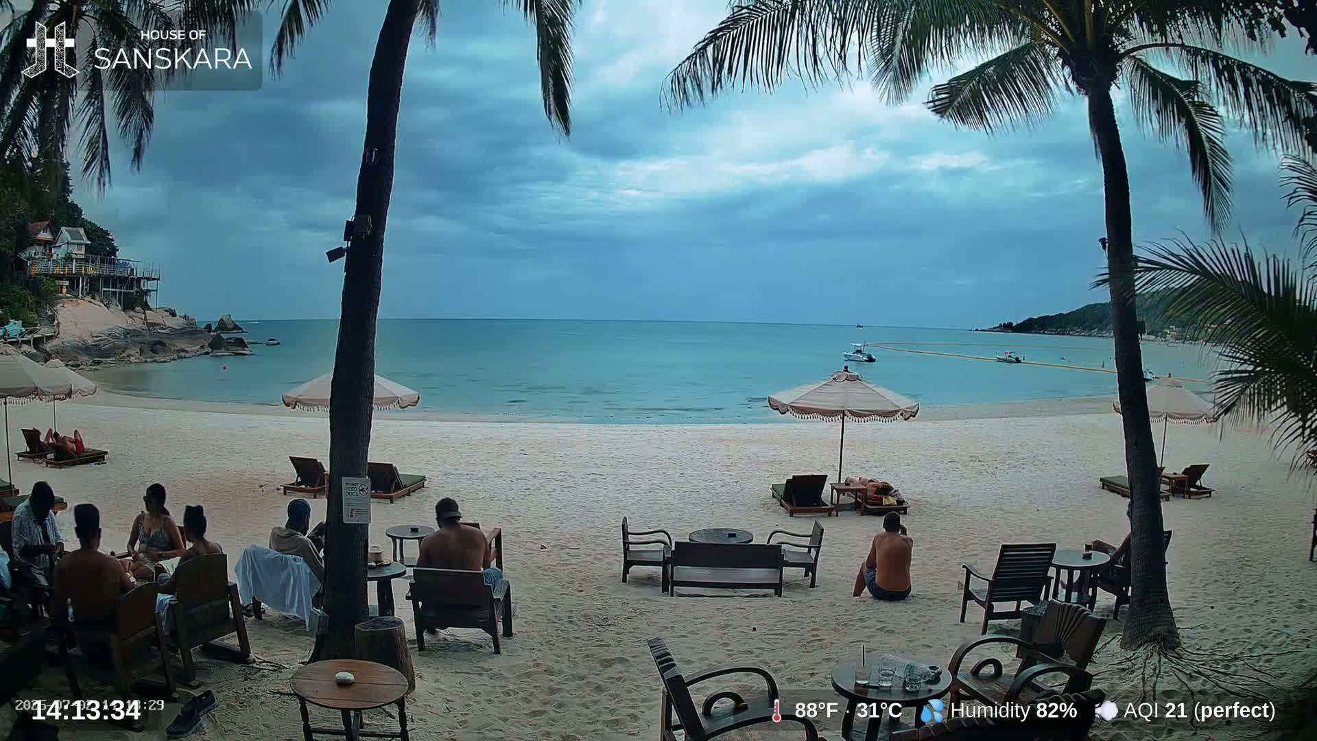 A sandy beach with people sitting at outdoor tables under umbrellas, overlooking a calm ocean under a partly cloudy sky.