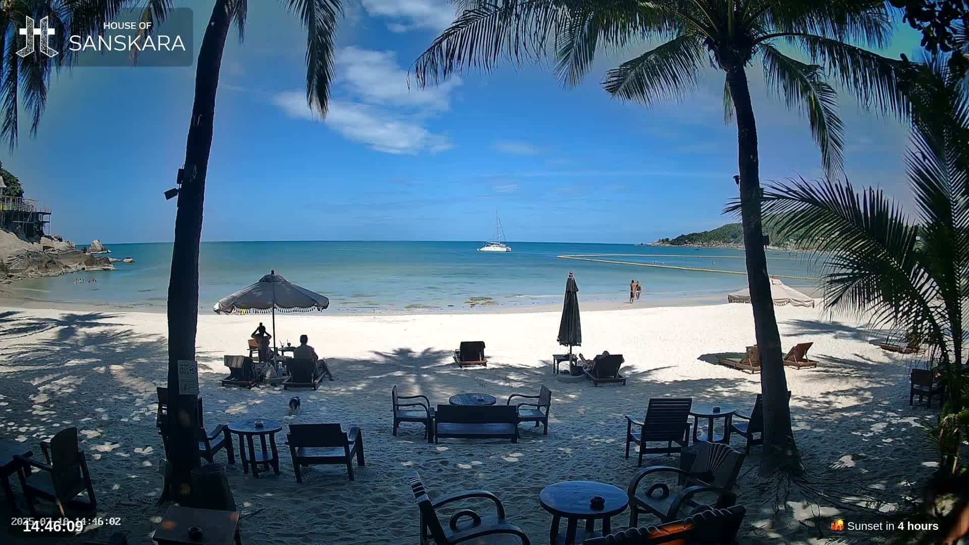 A sunny beach scene with calm, clear water, palm trees, and outdoor furniture situated on the white sand, with a sailboat visible in the distance.