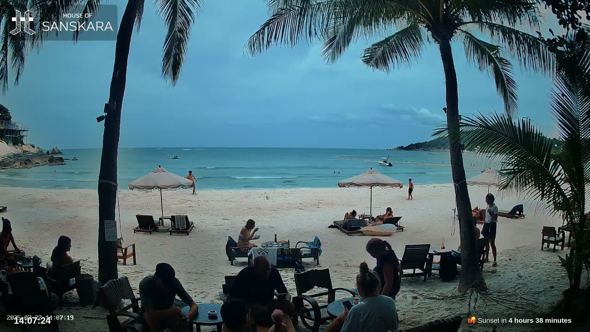 A beach bar on a sandy beach with people relaxing under umbrellas, palm trees, and an overcast sky.