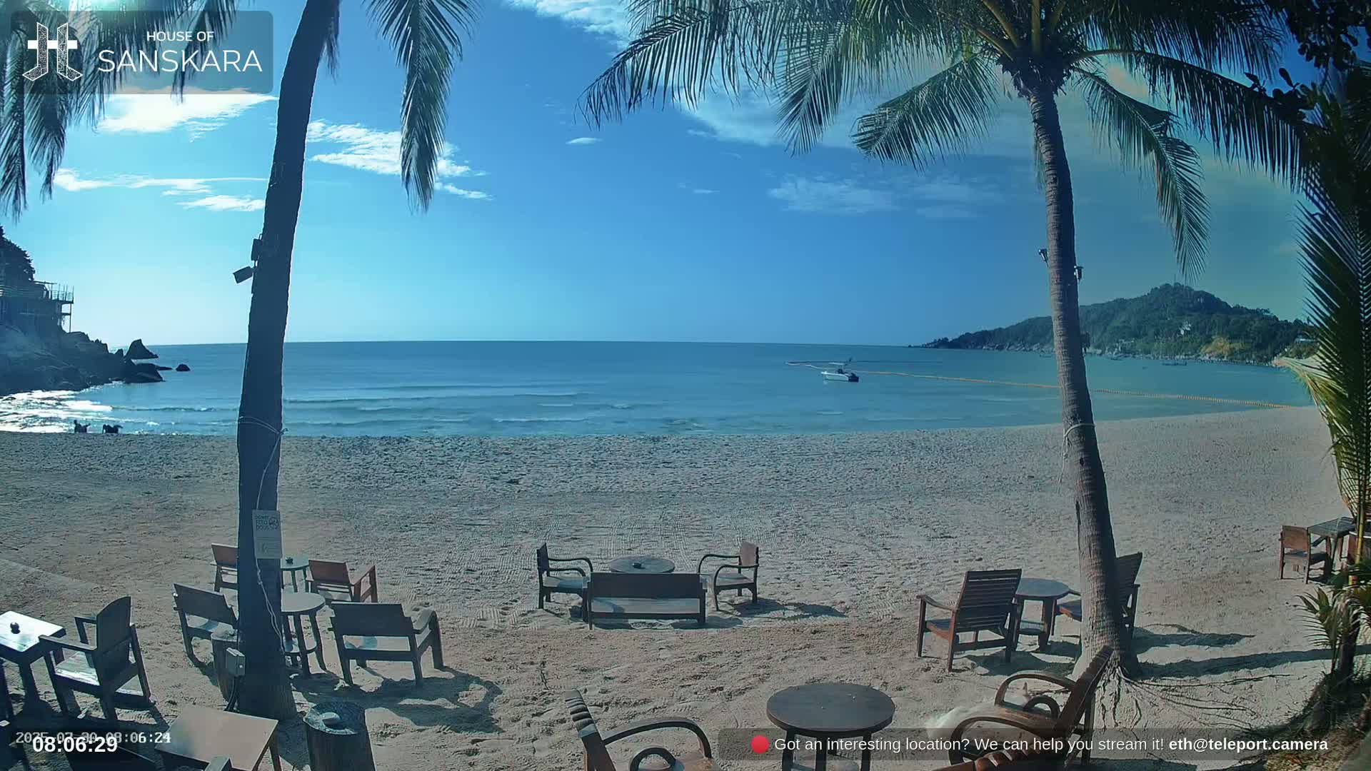 A beach bar on a sandy beach with people relaxing under umbrellas, palm trees, and an overcast sky.