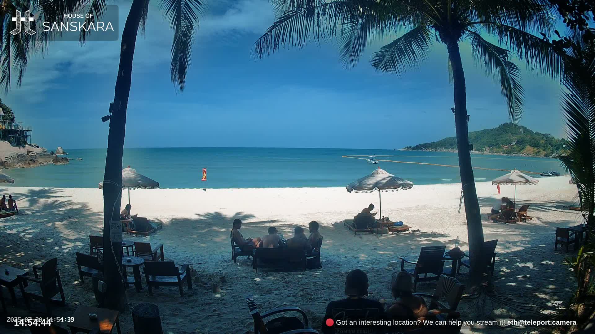 A beach scene on a sunny day shows a white sandy beach with turquoise water, palm trees, beach umbrellas, and people relaxing on lounge chairs and at outdoor tables.