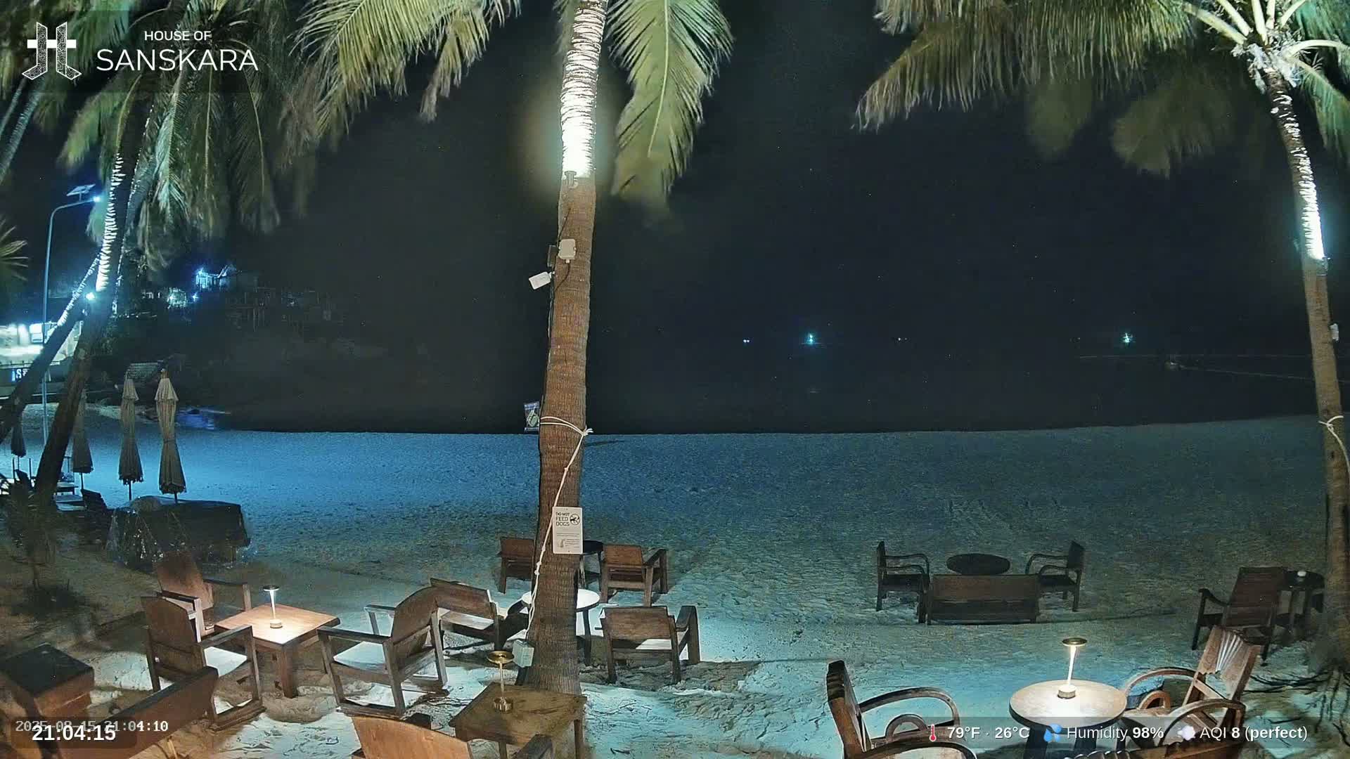 A tranquil beach scene at twilight shows a calm ocean with gentle waves lapping the shore, several empty tables and chairs on the sand, and palm trees silhouetted against the sky.