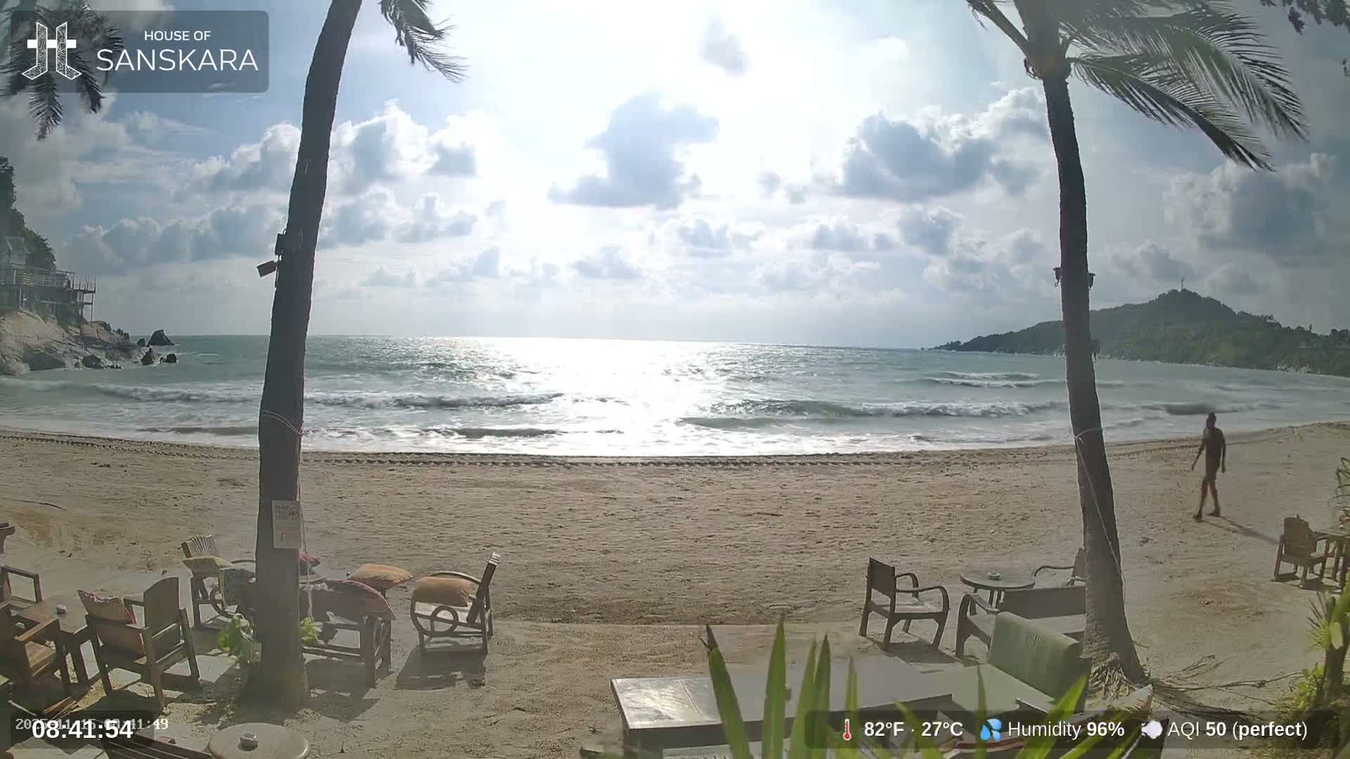 A bright, partly cloudy day illuminates a sandy beach with crashing waves, two prominent palm trees framing the view, outdoor seating, and a distant figure walking along the shore.