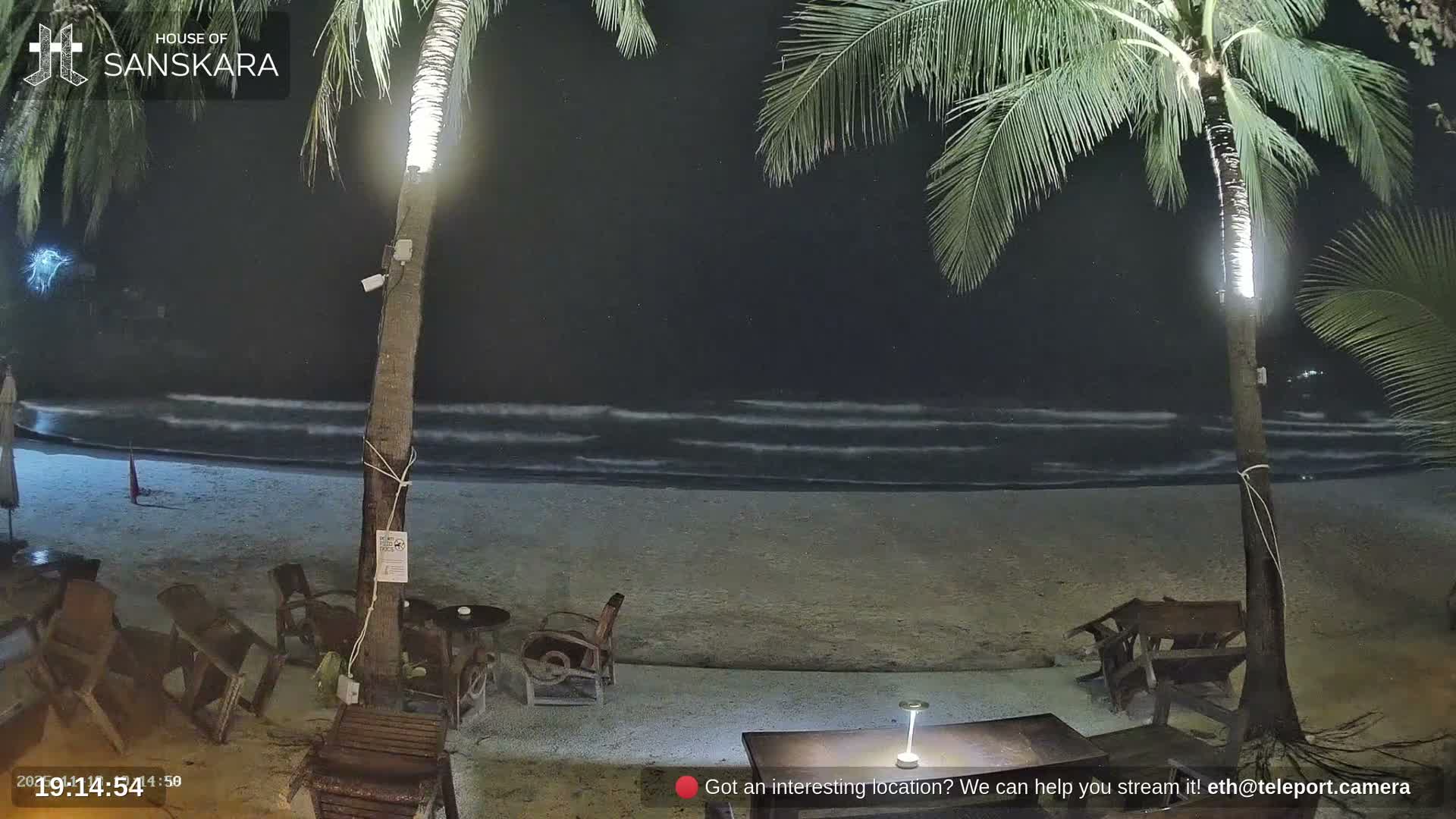 A bright, partly cloudy day illuminates a sandy beach with crashing waves, two prominent palm trees framing the view, outdoor seating, and a distant figure walking along the shore.