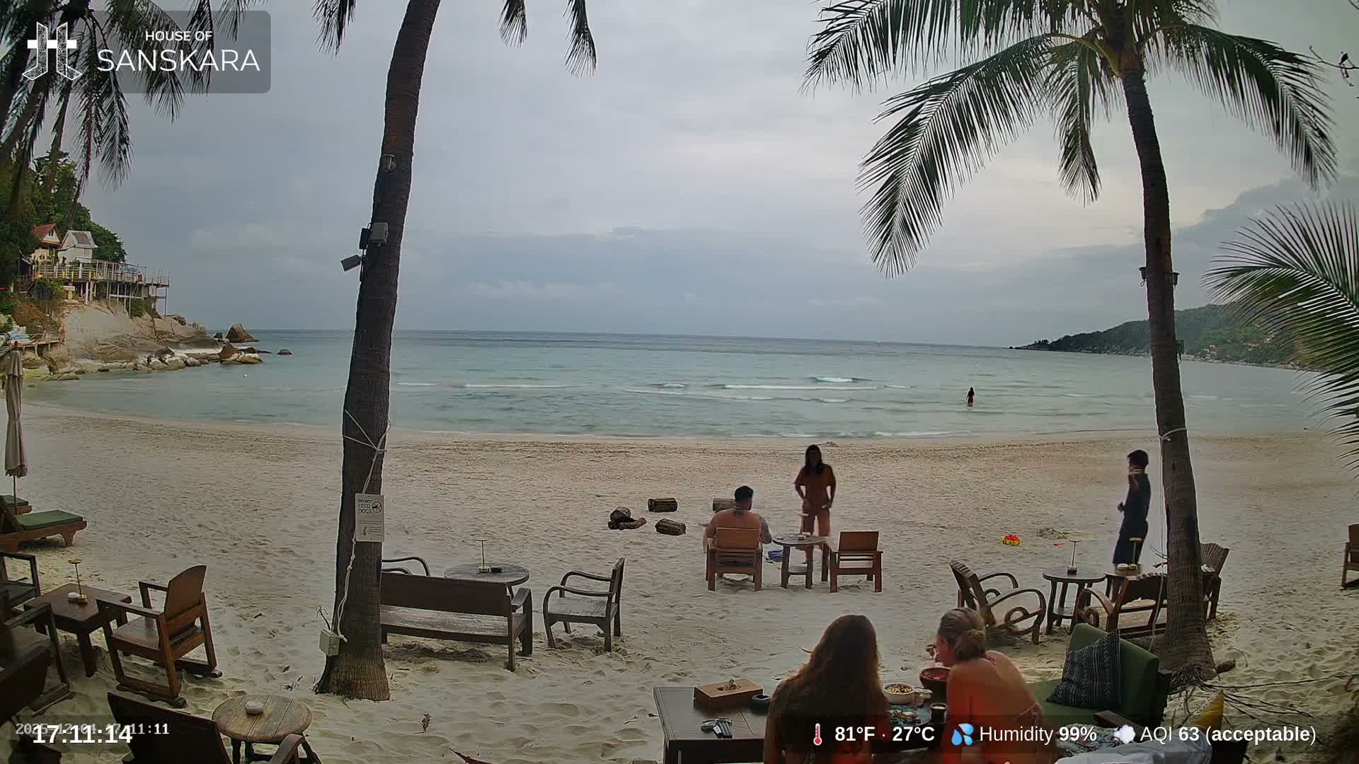 A cloudy day at a tropical beach resort shows people relaxing on the sandy shore, some seated at tables while others are near or in the calm ocean, framed by tall palm trees.