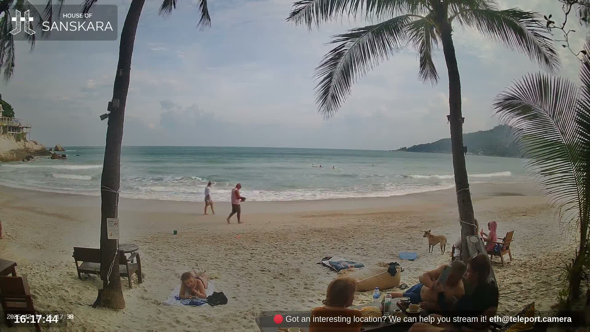 A cloudy day at a tropical beach resort shows people relaxing on the sandy shore, some seated at tables while others are near or in the calm ocean, framed by tall palm trees.