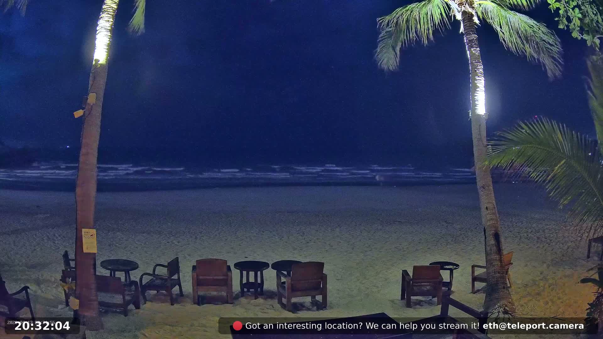 A cloudy day at a tropical beach resort shows people relaxing on the sandy shore, some seated at tables while others are near or in the calm ocean, framed by tall palm trees.