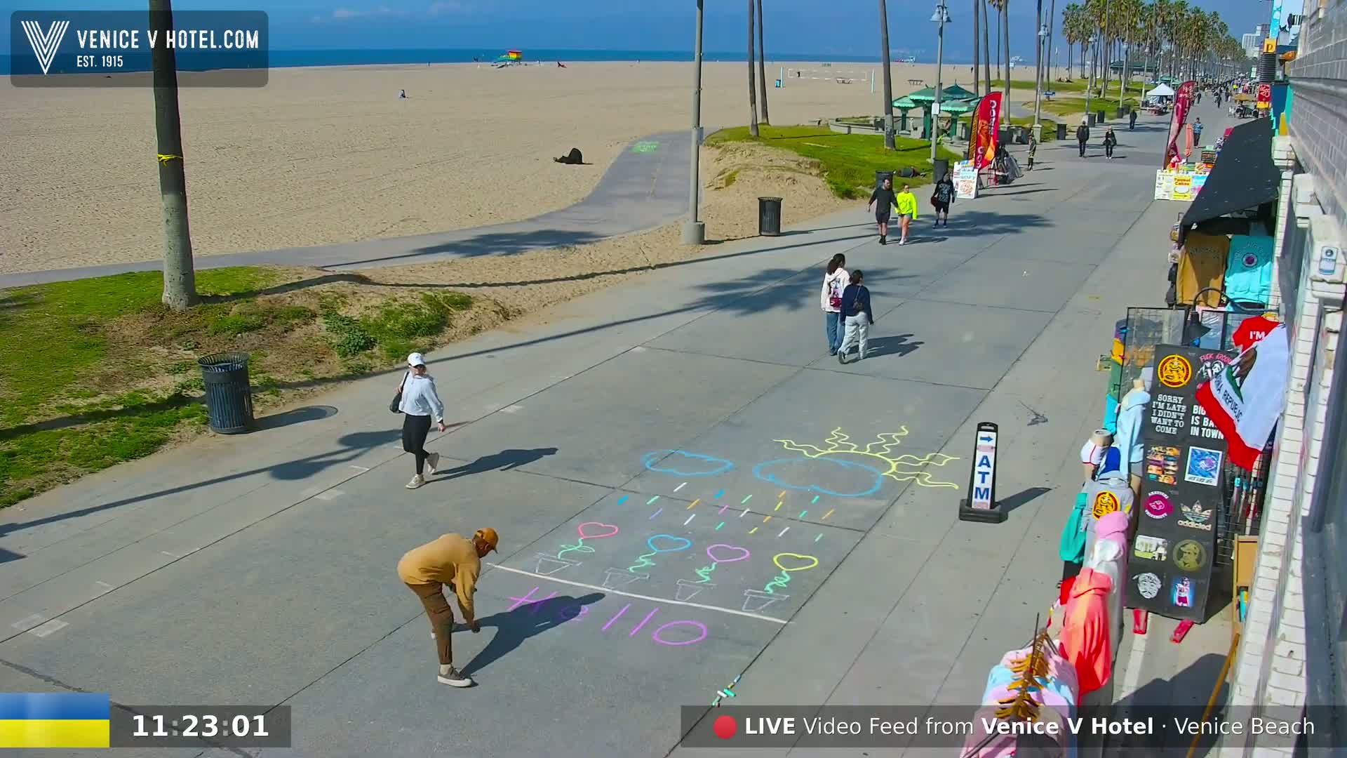A sunny beach boardwalk scene shows several people walking, a person drawing chalk art on the pavement, and beach shops.