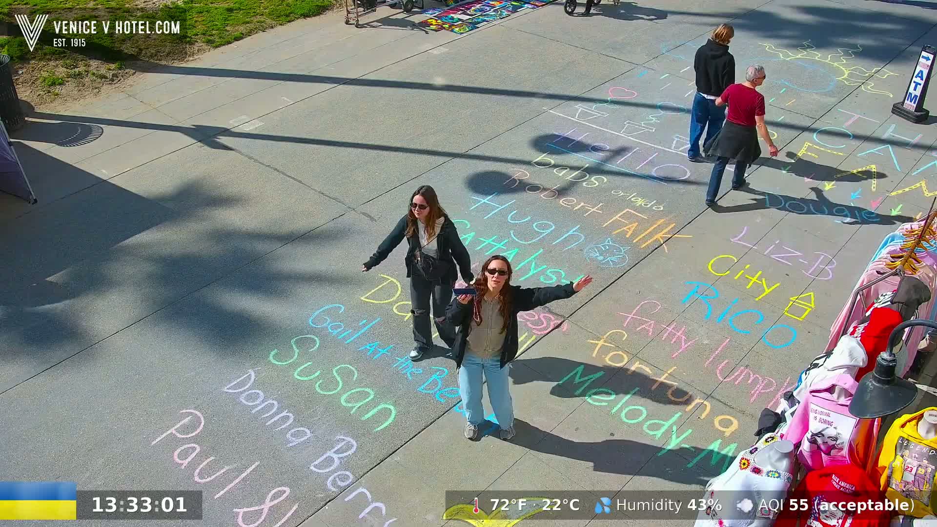 Two women stand on a sidewalk with numerous names written in chalk on the ground, under sunny conditions.
