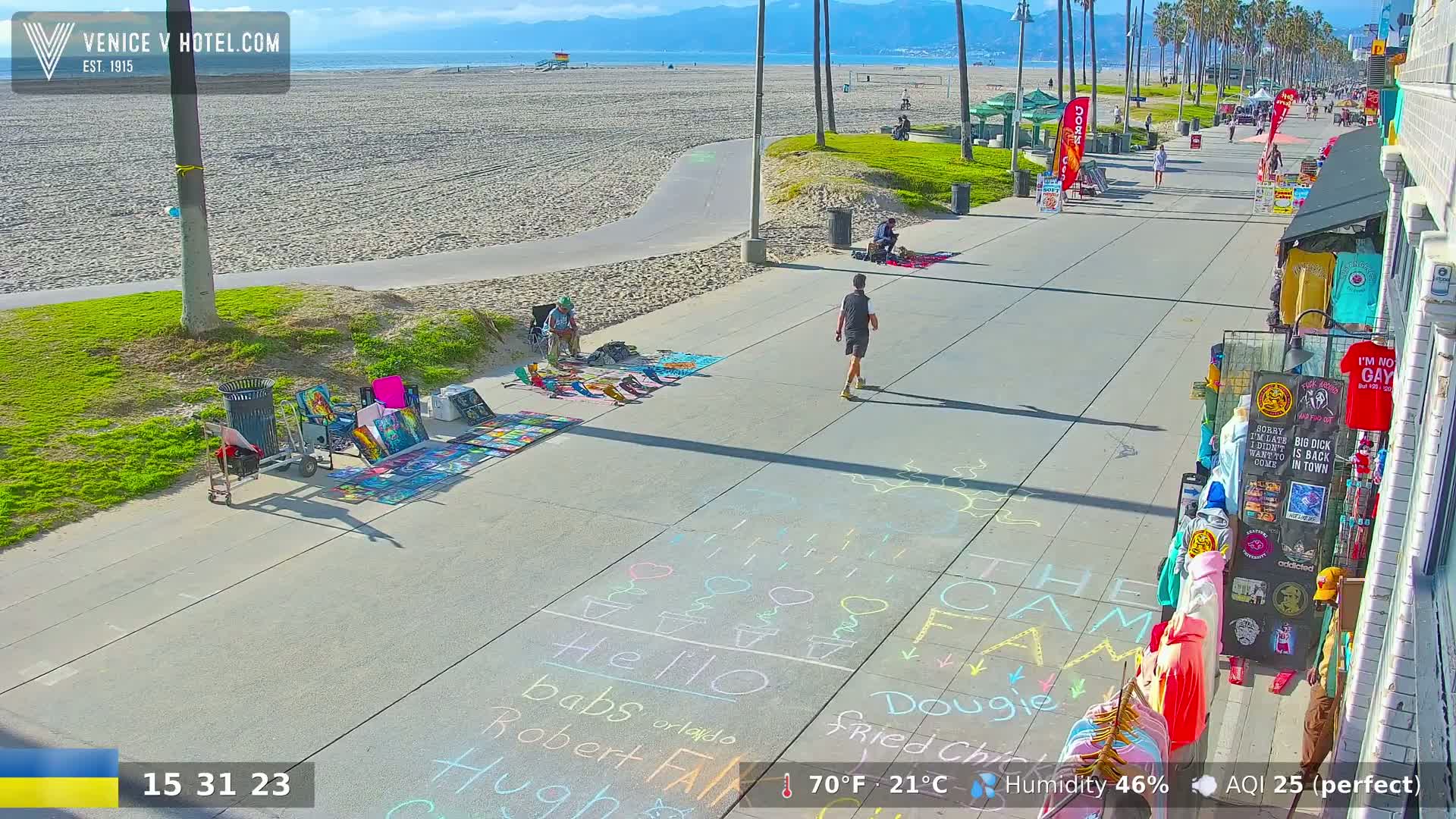 A paved walkway next to a sandy beach shows several people walking and artists displaying artwork; the weather is sunny and clear.