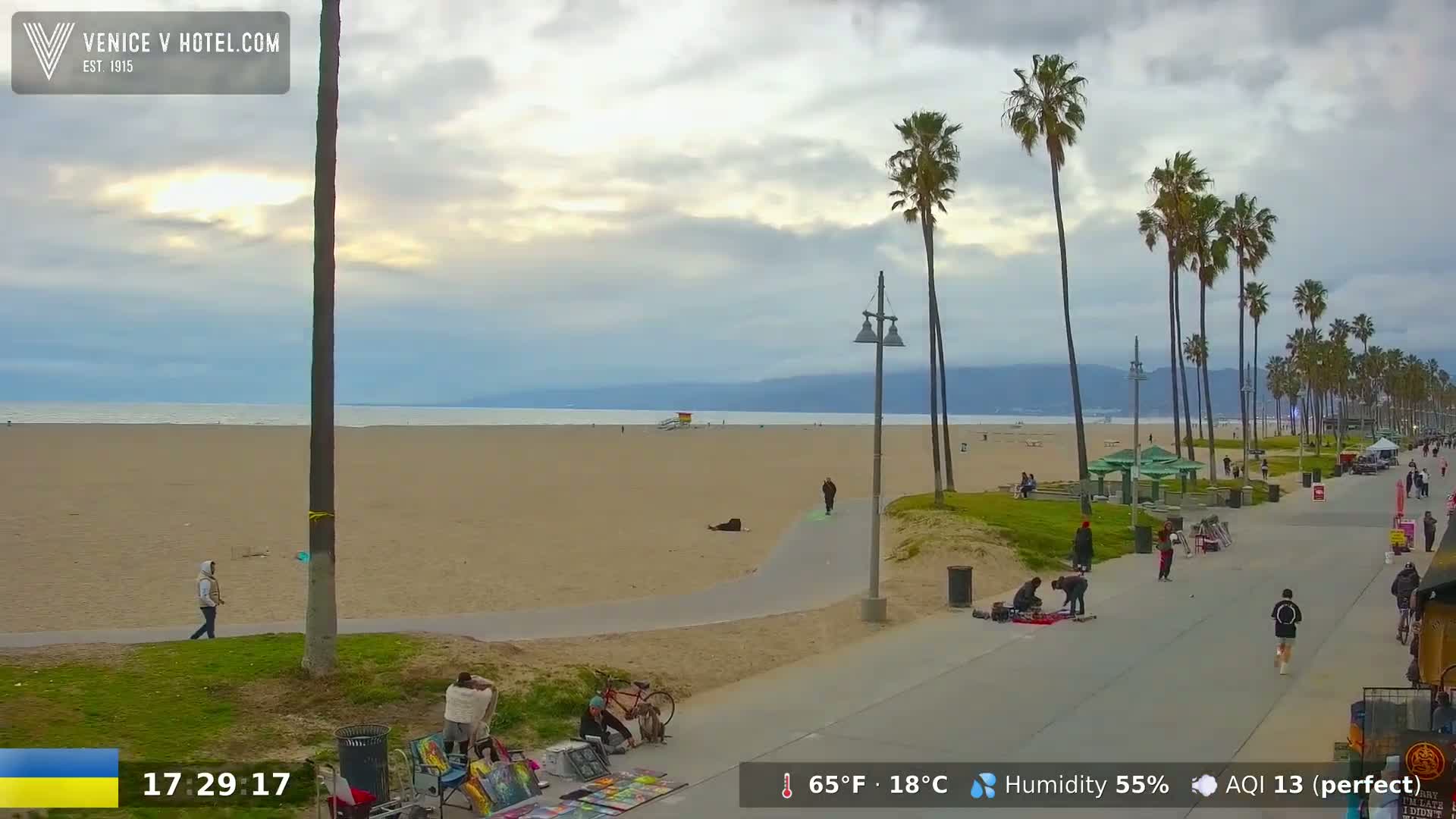 A mostly cloudy day at a beach with palm trees lining a boardwalk, where several people are walking and some are sitting on the sand or selling artwork.