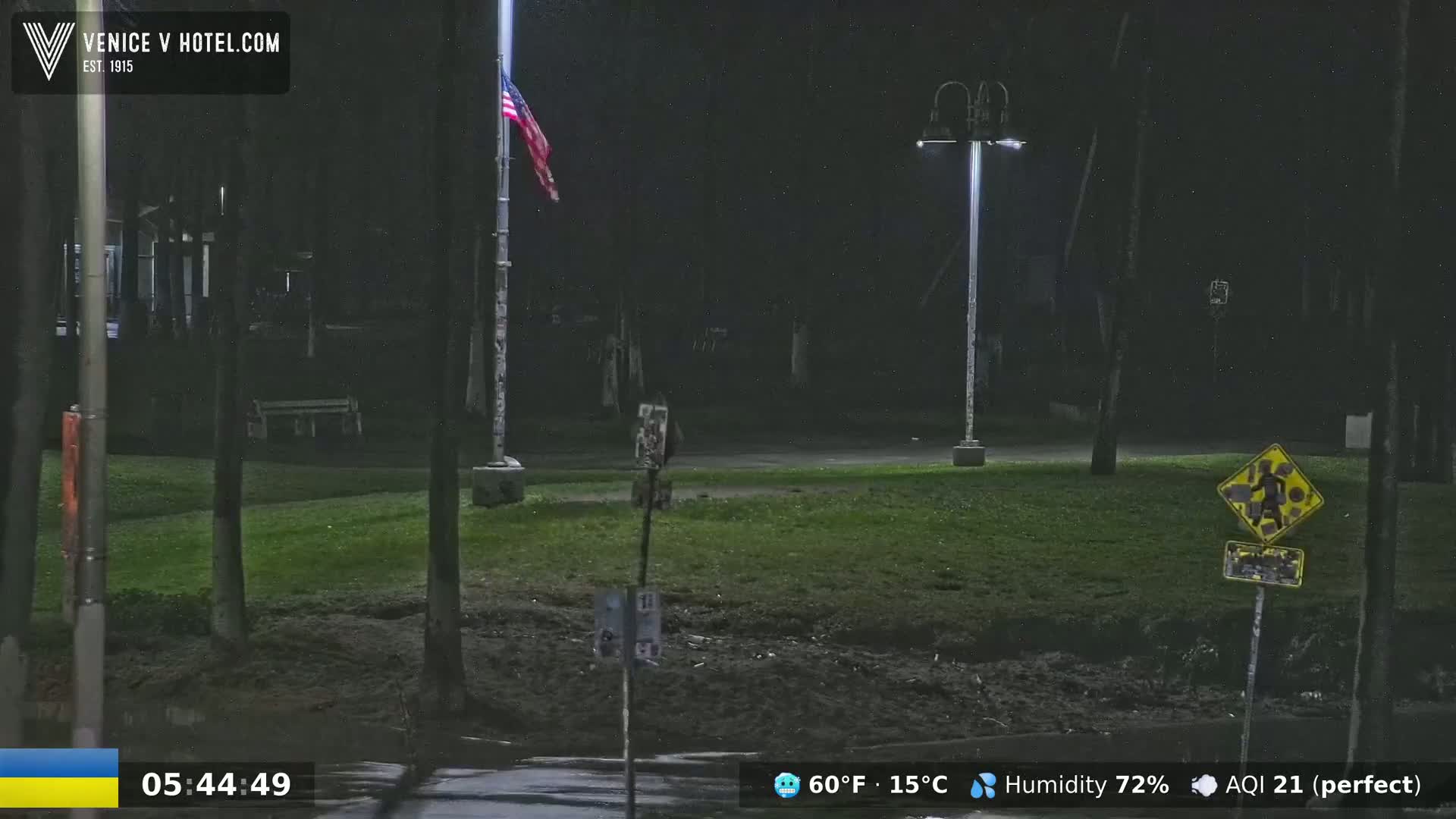 A nighttime view of a grassy area with a flagpole, streetlights, and a pedestrian crossing sign, under a dark sky with a temperature of 60°F (15°C) and 72% humidity.