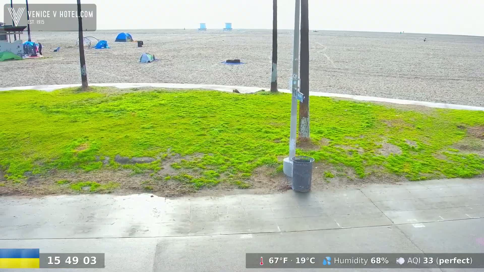 A mostly overcast day shows a patch of bright green grass next to a gray sidewalk bordering a sandy beach with a few tents and people visible in the distance.
