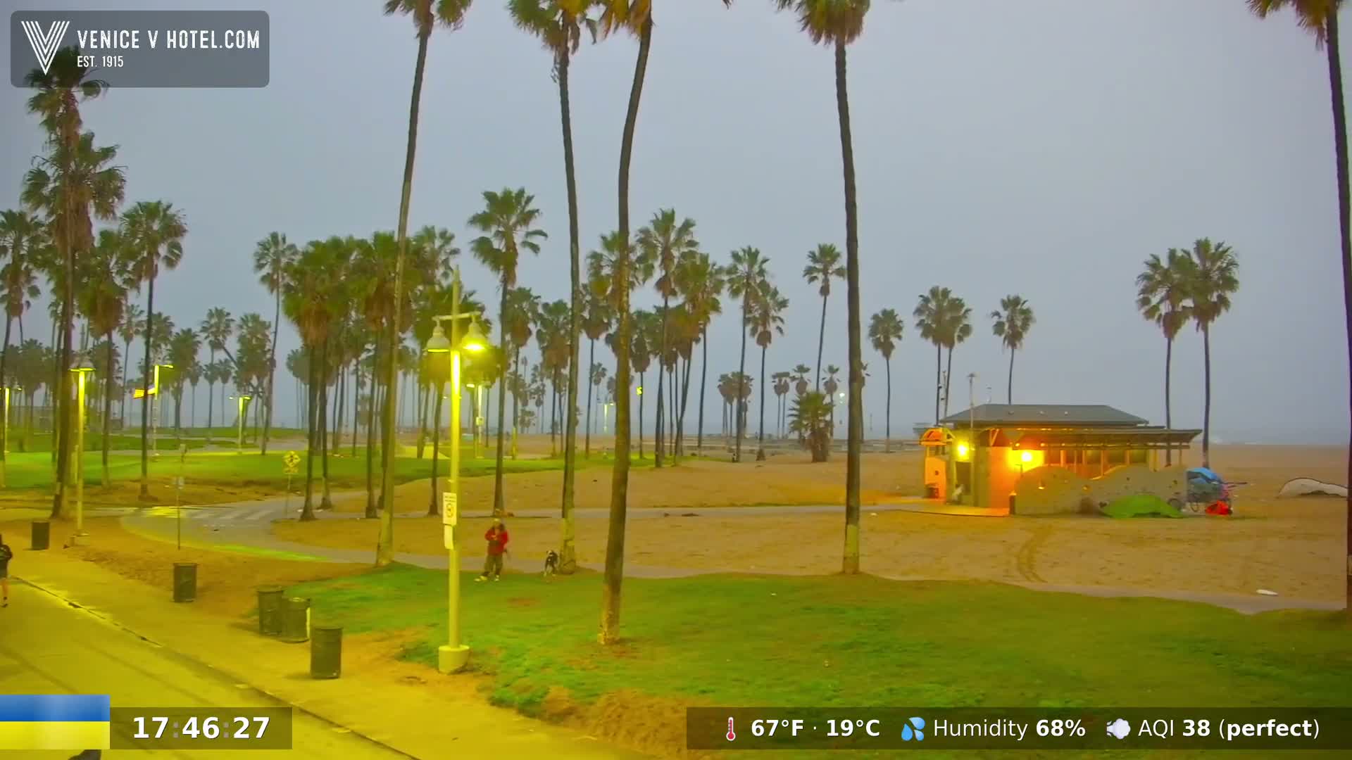 A beach scene at dusk shows a sandy beach, palm trees, a small building, and a paved walkway under a hazy, overcast sky.