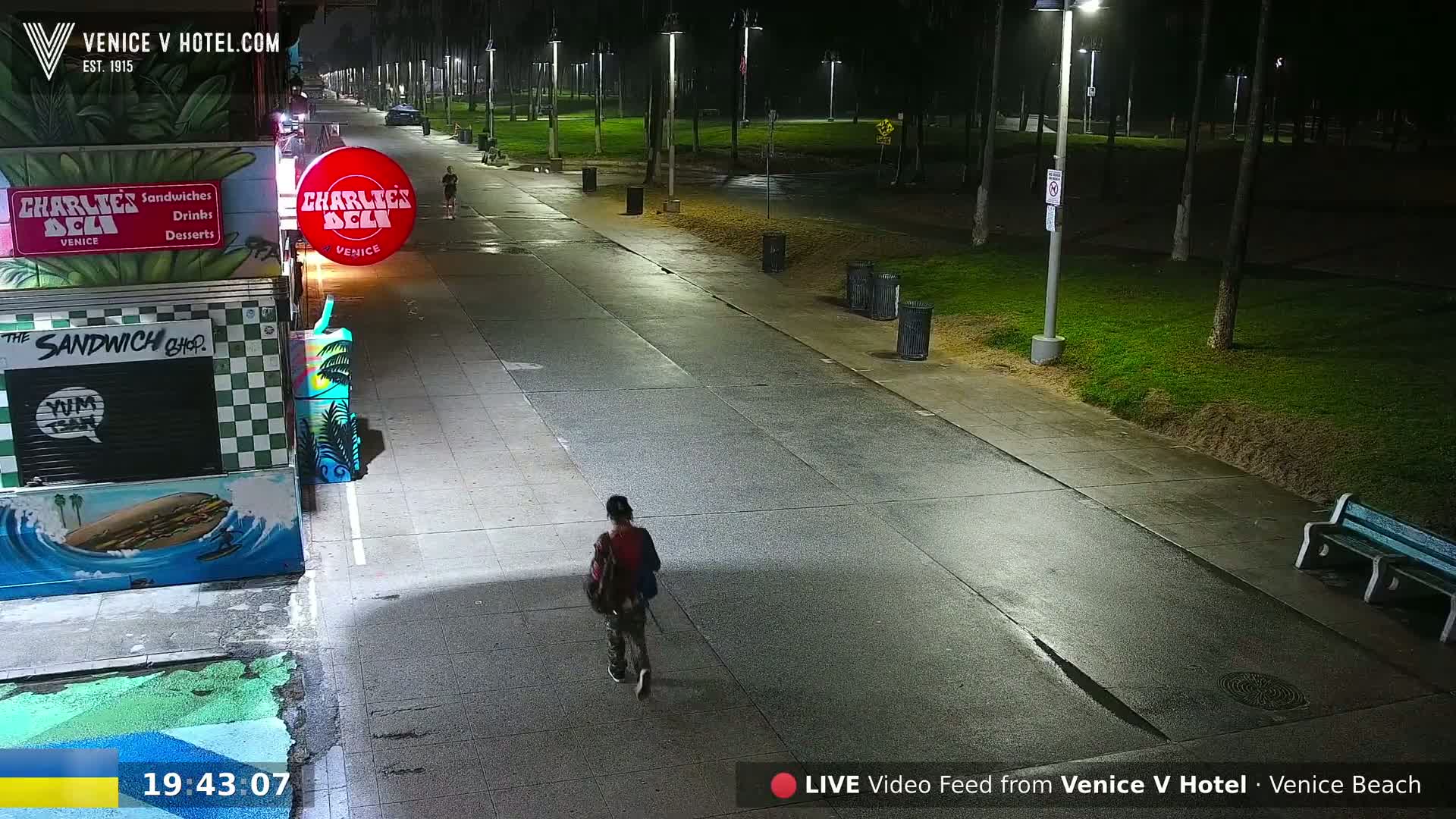 A person walks down a wet, nighttime boardwalk beside a park, passing a brightly-lit deli.