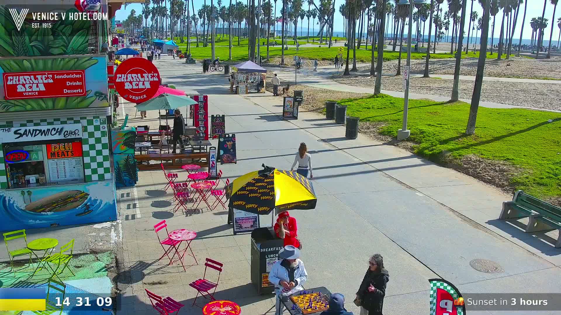 A sunny boardwalk scene shows people walking, sitting at outdoor tables, and playing chess near food stands under palm trees next to a beach.