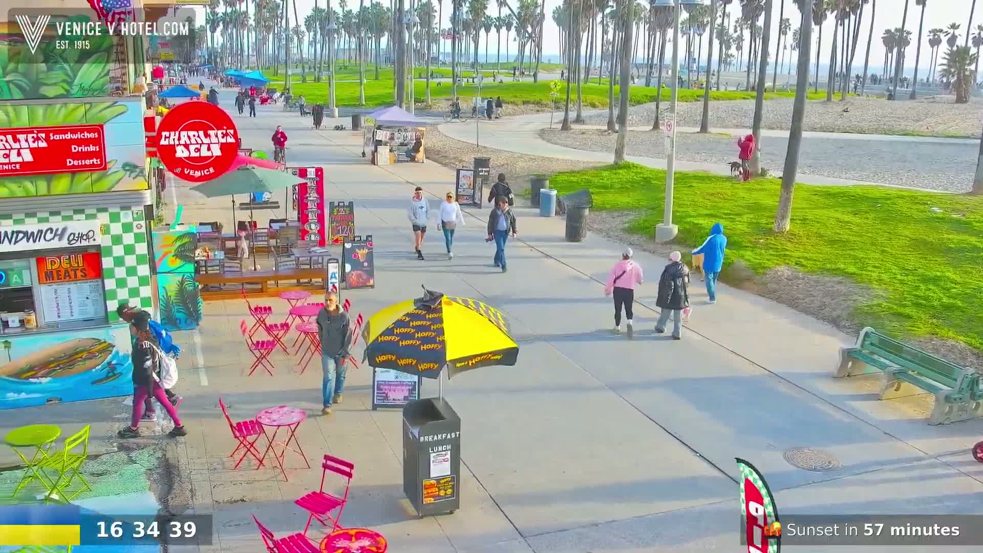 A sunny boardwalk scene shows several people walking past outdoor eateries and a beach area with palm trees.