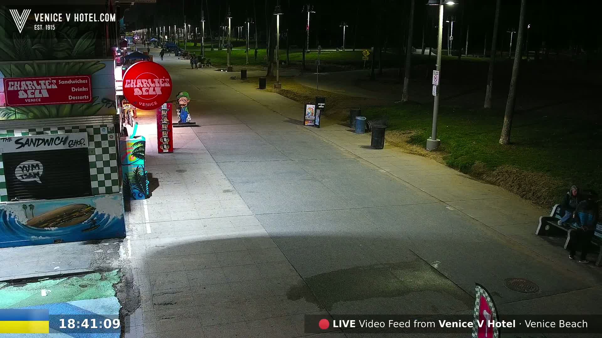 A nighttime view of a paved walkway, lined with streetlights and bordering a grassy area, shows a few people sitting on a bench near some food stalls.