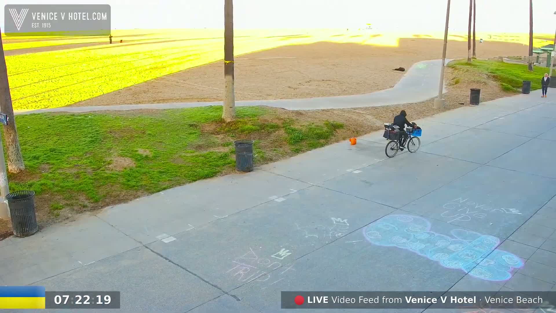 A person rides a bicycle on a paved path next to a beach, with chalk drawings on the ground and a mostly sunny sky.