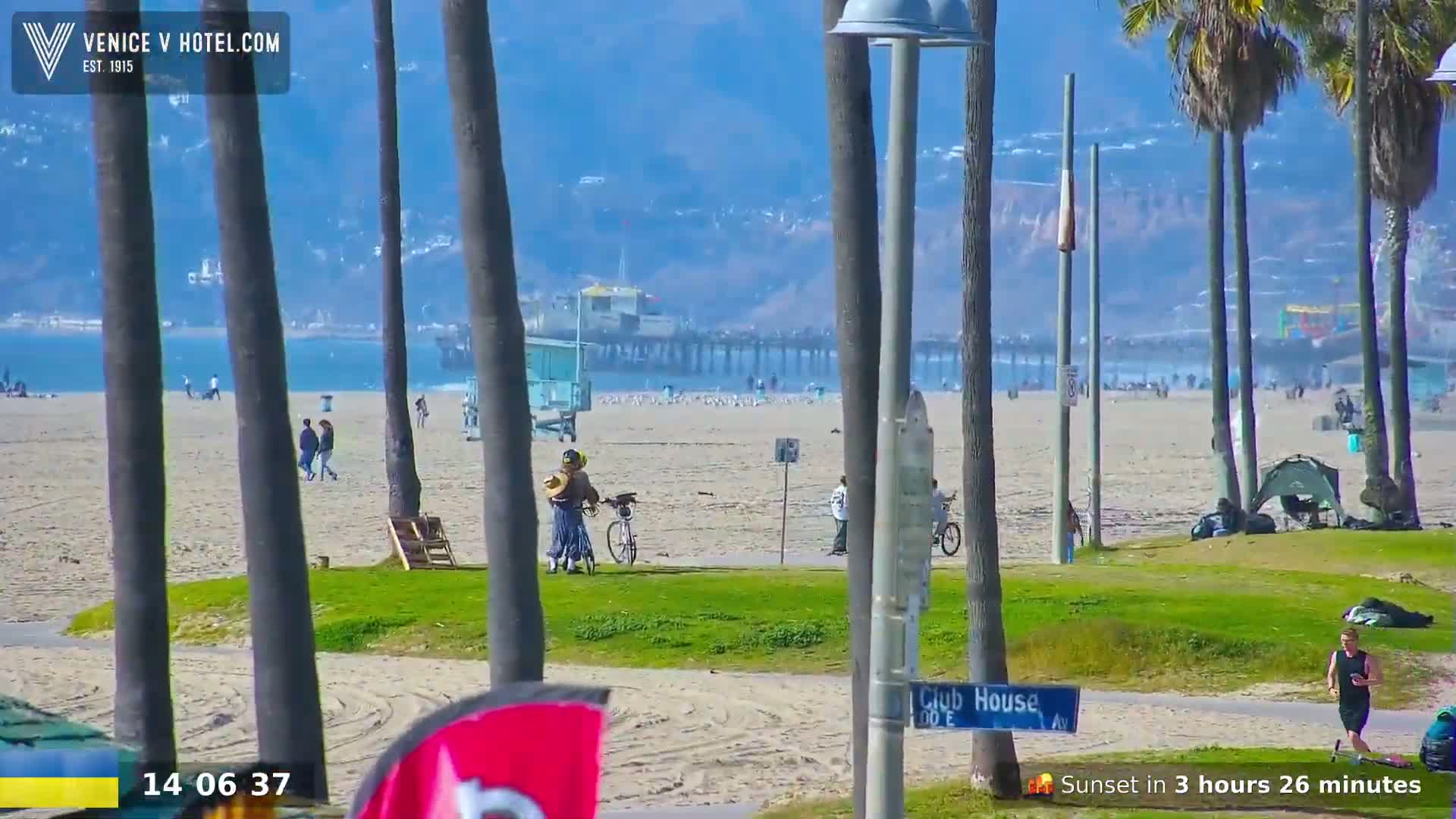 A sunny beach scene shows a pier in the distance, people walking on the sand and grass, palm trees lining the beachfront, and a person with a bicycle.