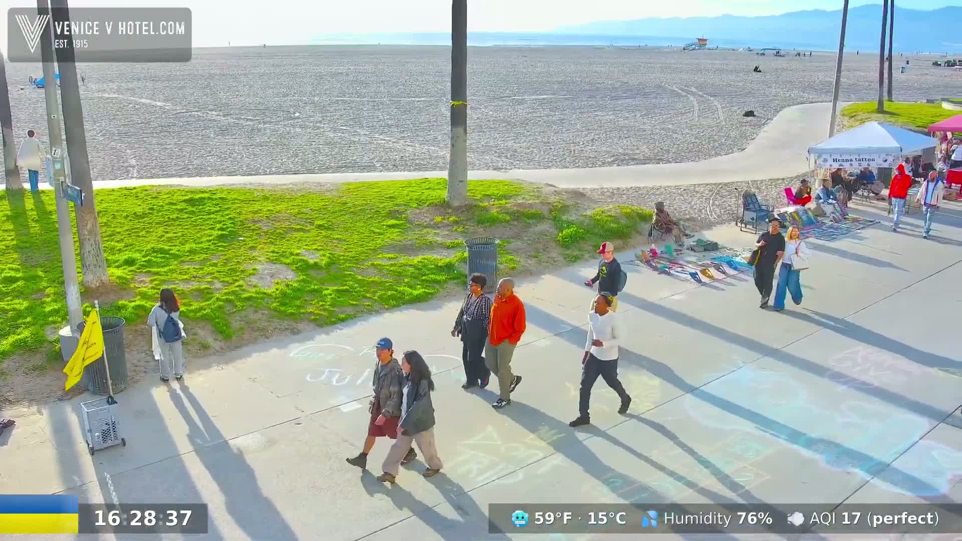 A sunny day at a beach boardwalk shows several people walking along a paved path beside a sandy beach and grassy area, with various vendors set up along the path.