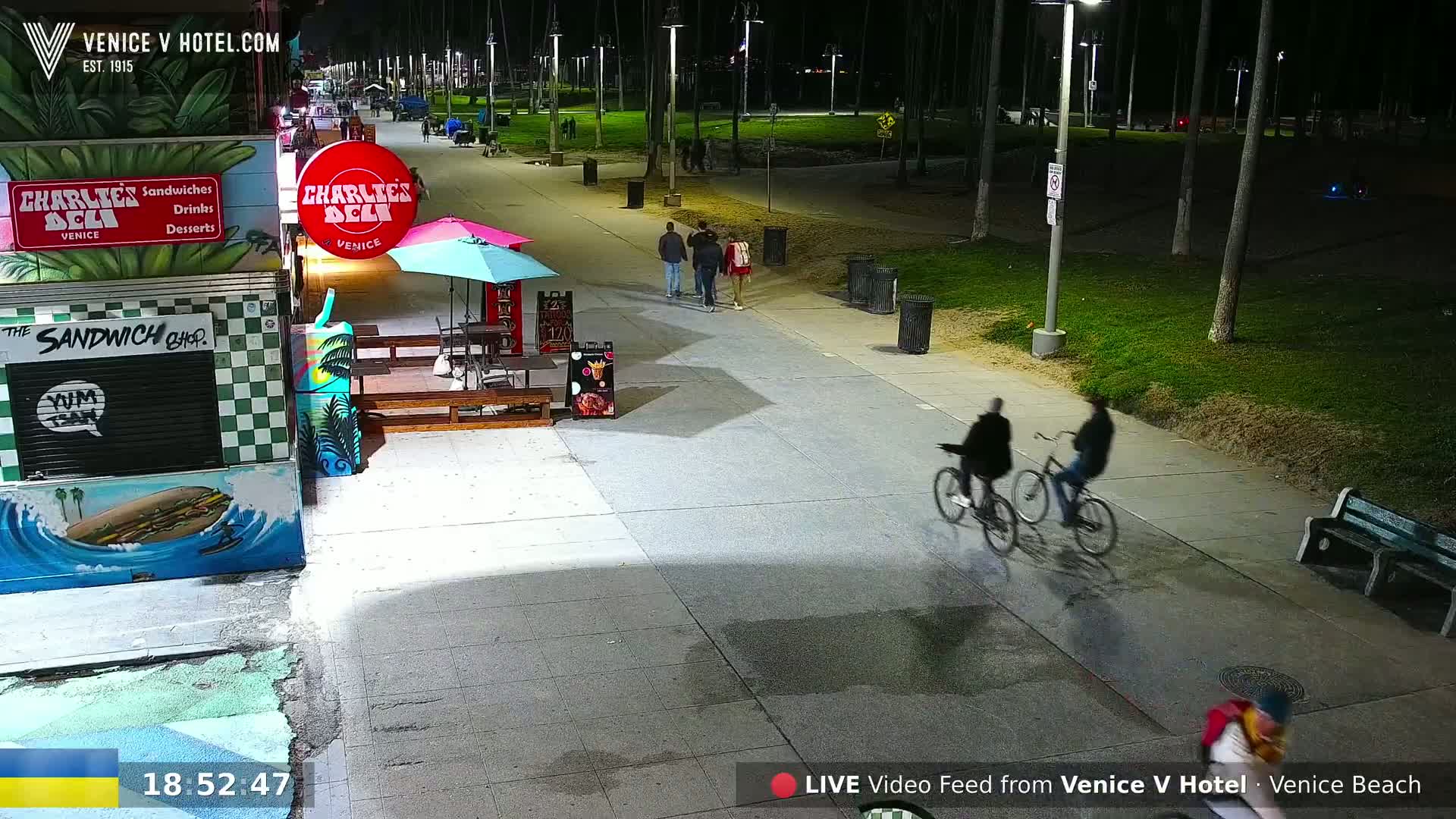At night, several people walk and bicycle along a paved beachfront path lined with palm trees and food stands.