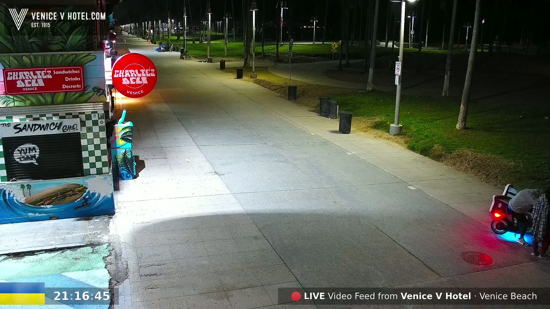 A nighttime view of a paved walkway next to a beachside park, with a food stand and a person on a motorized scooter visible.