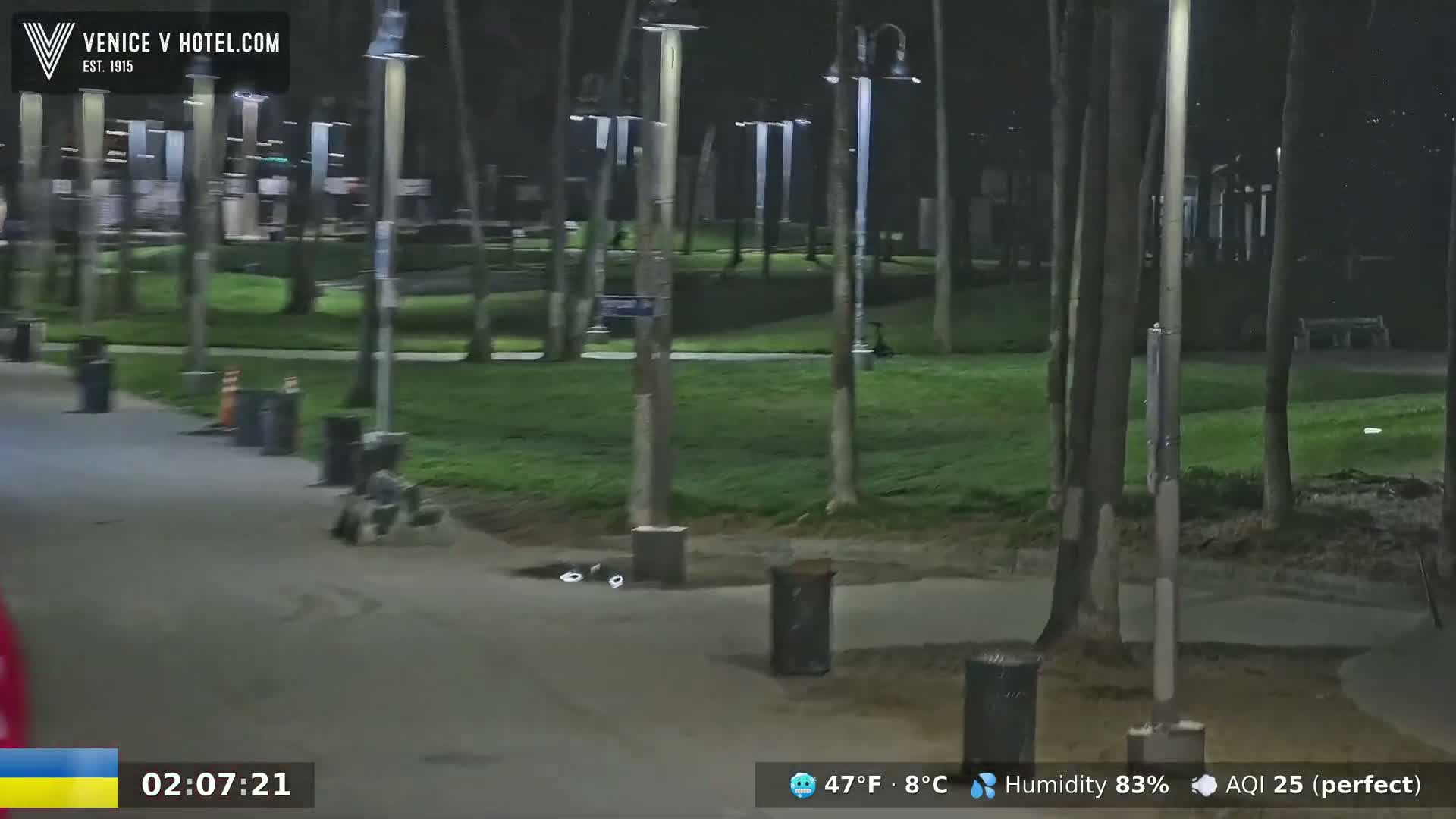 A nighttime view of a paved pathway through a park with trees and trash cans, under mostly clear skies at 47°F and 83% humidity.