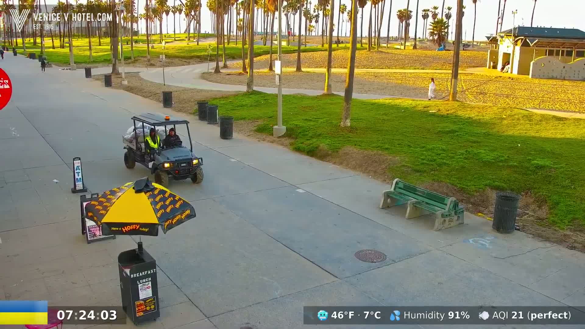 A golf cart with two people in it is parked near a food stand on a paved walkway beside a grassy area with palm trees, a beach, and a building in the background, under sunny skies.
