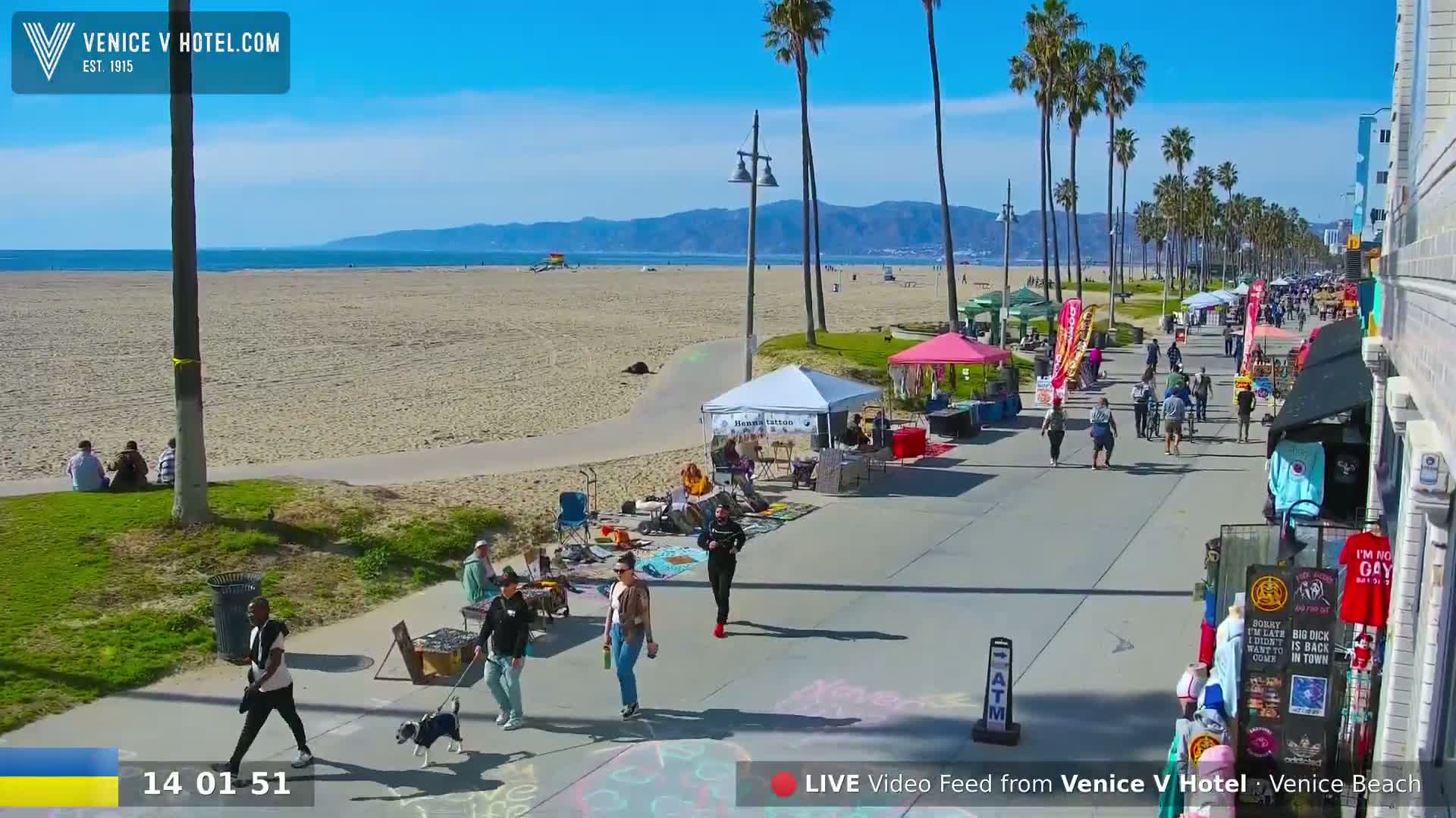 A sunny day at Venice Beach shows a boardwalk with vendors and people walking, leading to a sandy beach and ocean under a clear blue sky.
