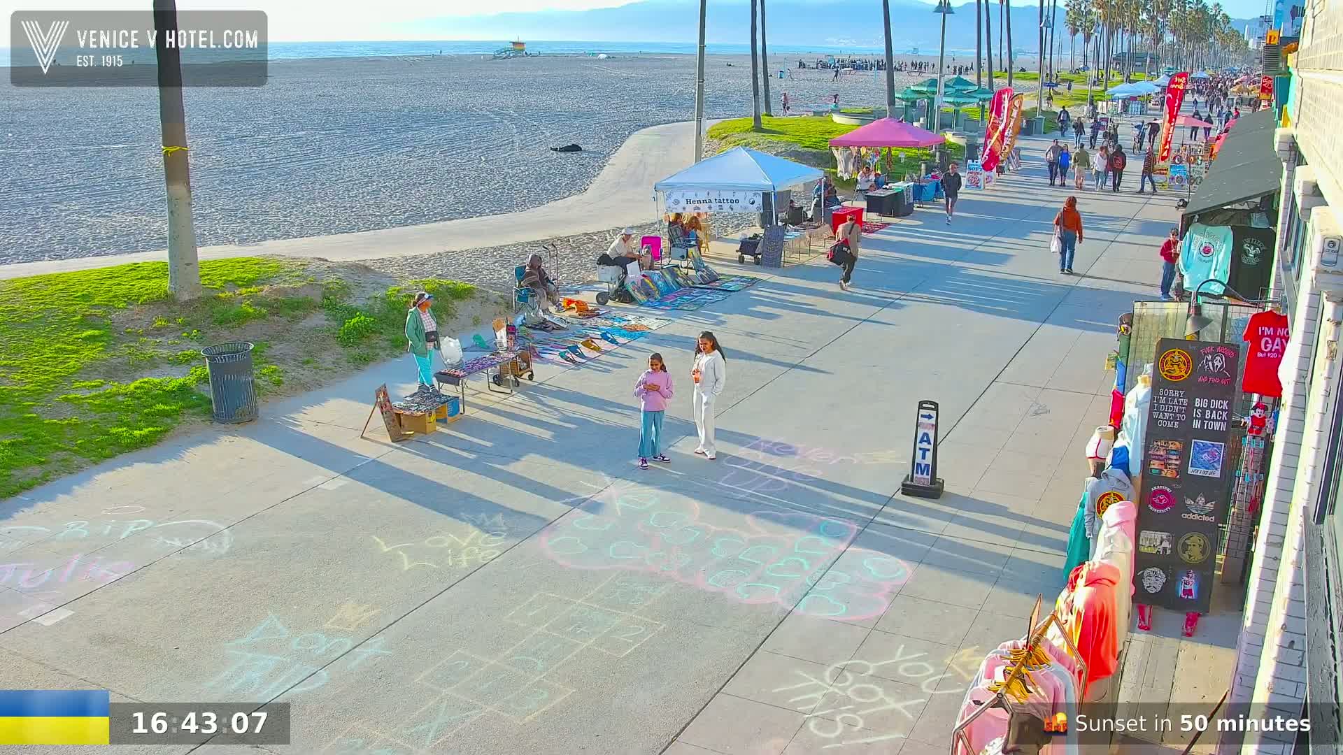 A sunny beachfront boardwalk scene shows various vendors selling goods, people walking along the promenade, and children playing hopscotch drawn in chalk on the pavement.